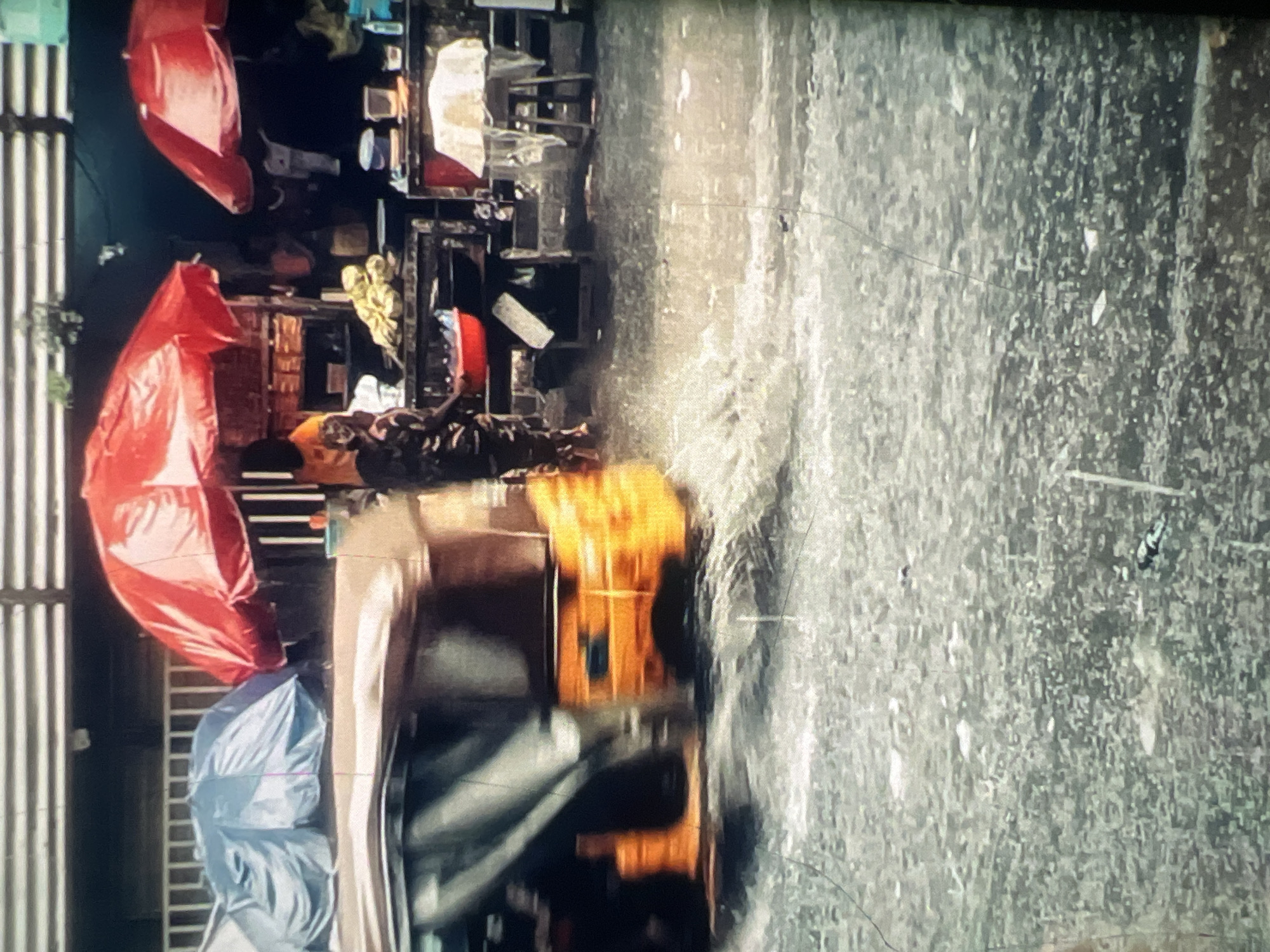 The image captures a street scene in Lagos, Nigeria, likely during or after a heavy rainfall. The ground is flooded with water, reflecting the surrounding environment. In the foreground, a blurred yellow and orange auto-rickshaw, also known as a "keke," is in motion, creating ripples and splashes in the water as it moves.
Behind the keke, a market stall is set up under large red umbrellas. There are people at the stall, one of whom appears to be a vendor, possibly selling produce like bananas, which are visible on a table. Another person is seen in the background. The market stalls are stocked with various goods, some of which are covered. A blue tarp covers items on a shelf behind the red umbrellas. The overall impression is one of a bustling, yet temporarily disrupted, urban marketplace.