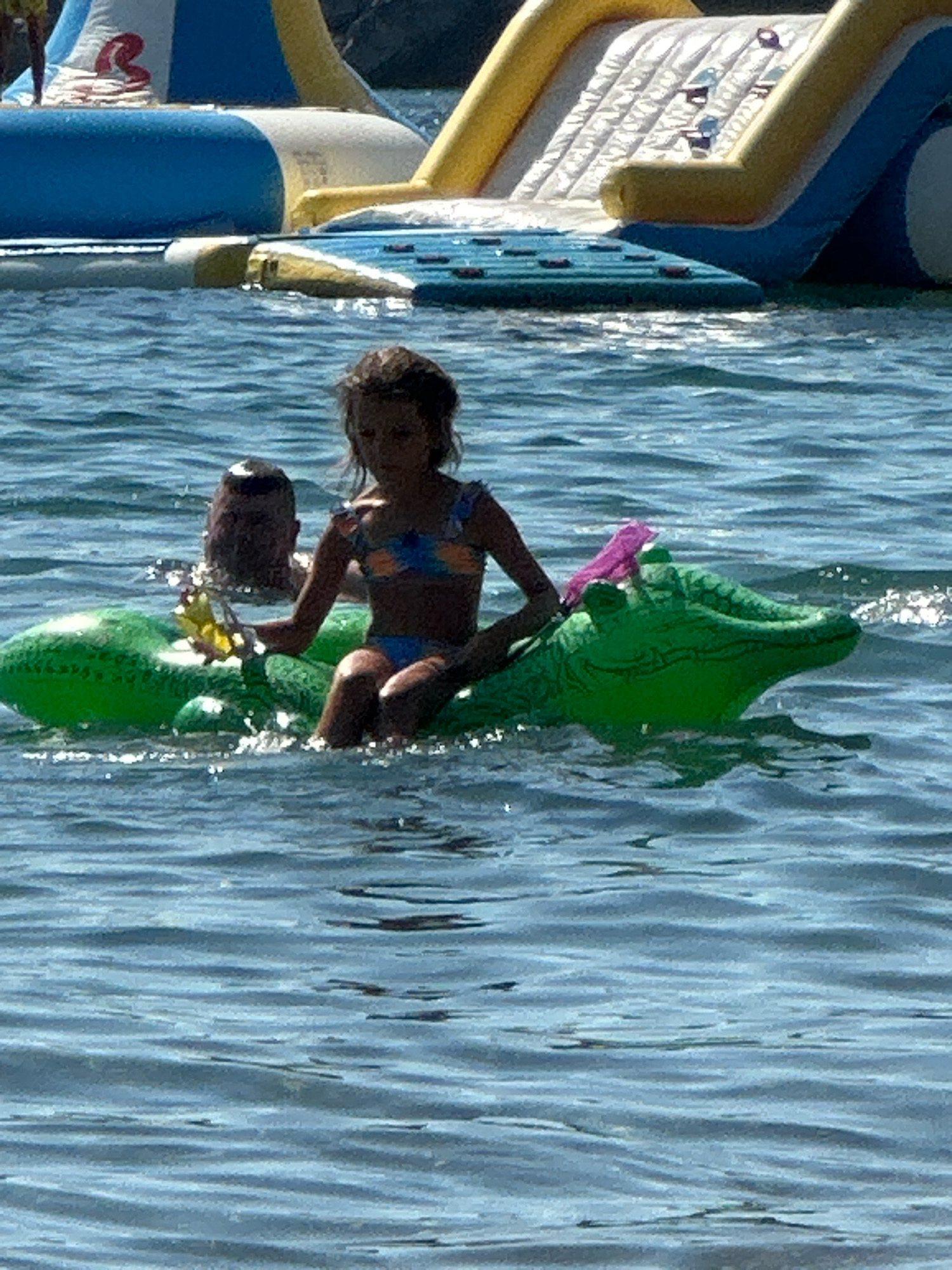 A young girl wearing a blue and orange bikini sits on an inflatable green alligator in the water. She holds a yellow flower in her left hand. Behind her, a person's head and shoulders are visible, partially submerged in the water. In the background, there is a large inflatable water park with blue, yellow, and white sections, including a slide. The water is choppy, suggesting a breezy day. The scene appears to be set during daylight hours. The girl's facial expression is not clearly discernible, but the overall impression is one of a fun day at the beach or a water park. The location is Boreti, Montenegro. There is no visible text in the image.