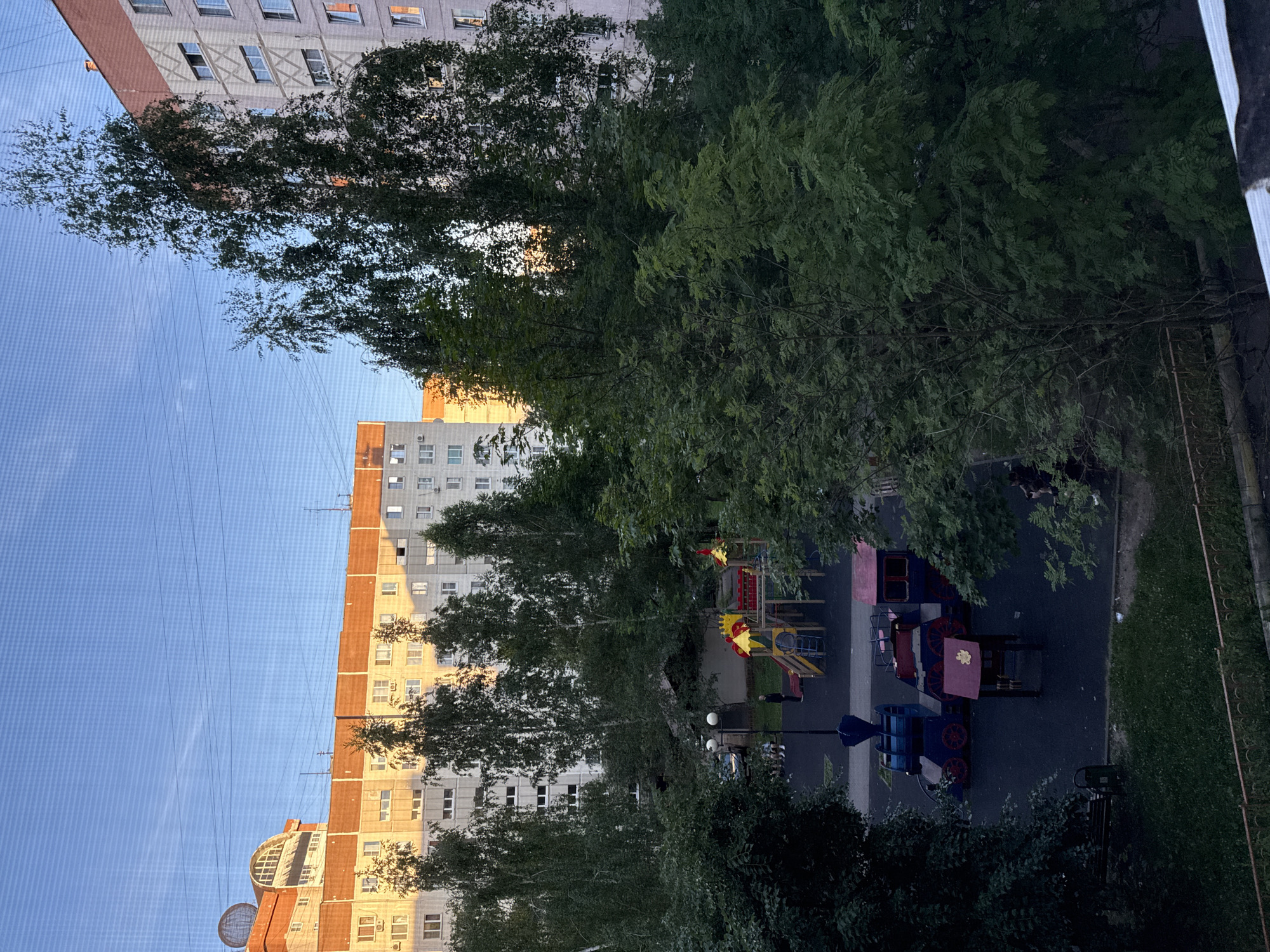 The image depicts a residential courtyard with a playground and apartment buildings under a clear sky. The playground features a multi-colored climbing structure with slides and a toy train set consisting of blue and red cars. Several trees, some with sparse foliage, are scattered throughout the scene, with dense green trees framing the foreground and mid-ground. Apartment buildings, some with brick accents and visible windows, line the background, illuminated by what appears to be late afternoon or early morning sunlight. A few figures are visible on or near the playground, and a bench is present in the lower left corner. The scene is viewed from an elevated position, possibly a window, with a fine mesh overlaying the image.