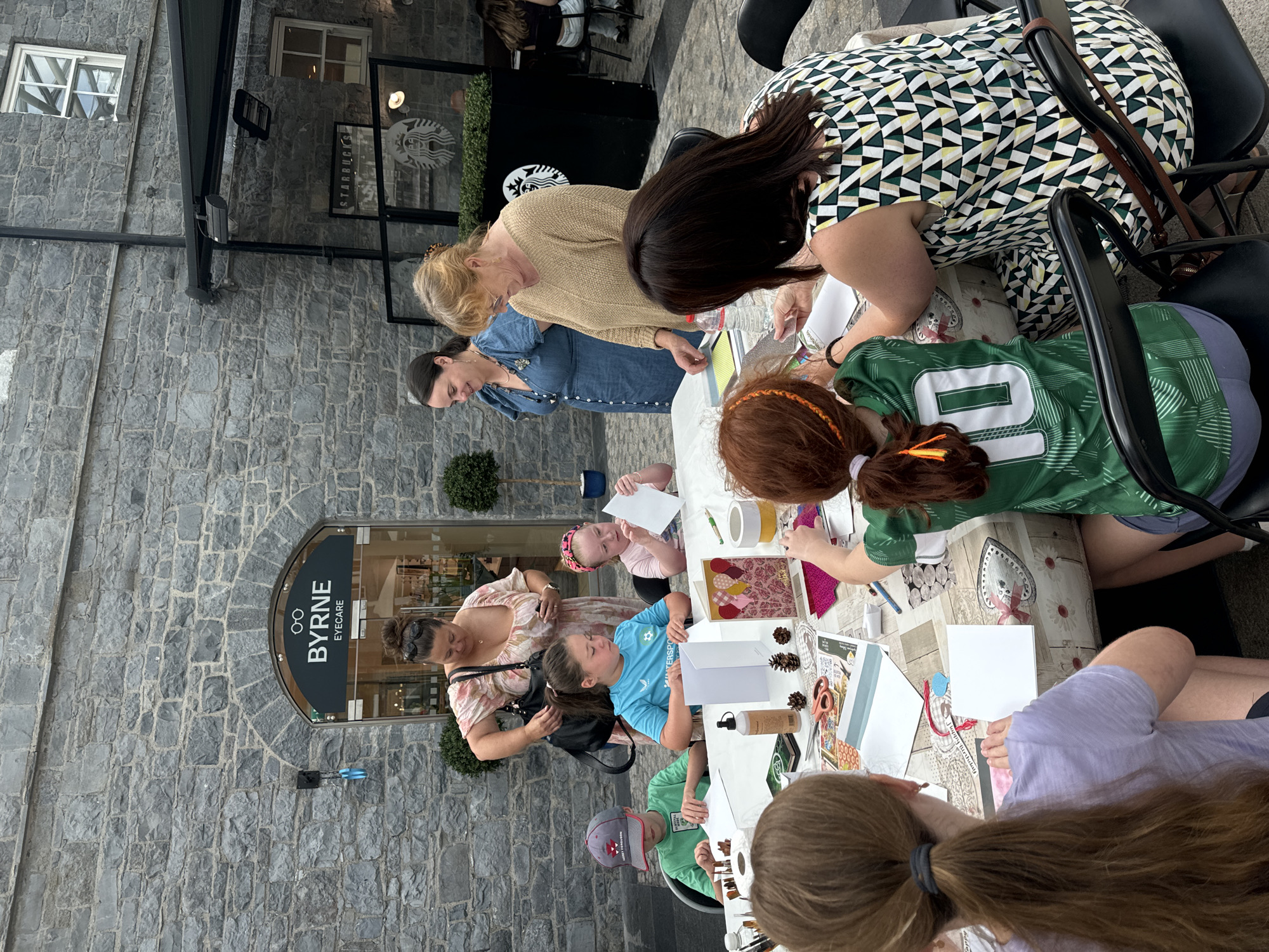 In Kilkenny, Ireland, a group of people are gathered at a craft table outdoors, in front of a building with a stone facade. A sign above an arched entrance reads "BYRNE EYECARE". Several individuals, including children and adults, are engaged in a crafting activity, with paper, scissors, glue, and decorative elements spread across the table. The scene appears to be set in a public area, possibly a courtyard or walkway, with a Starbucks visible in the background. The people are interacting, with some looking at their work and others conversing. The lighting suggests it is daytime, and the overall atmosphere is one of community engagement and creativity.