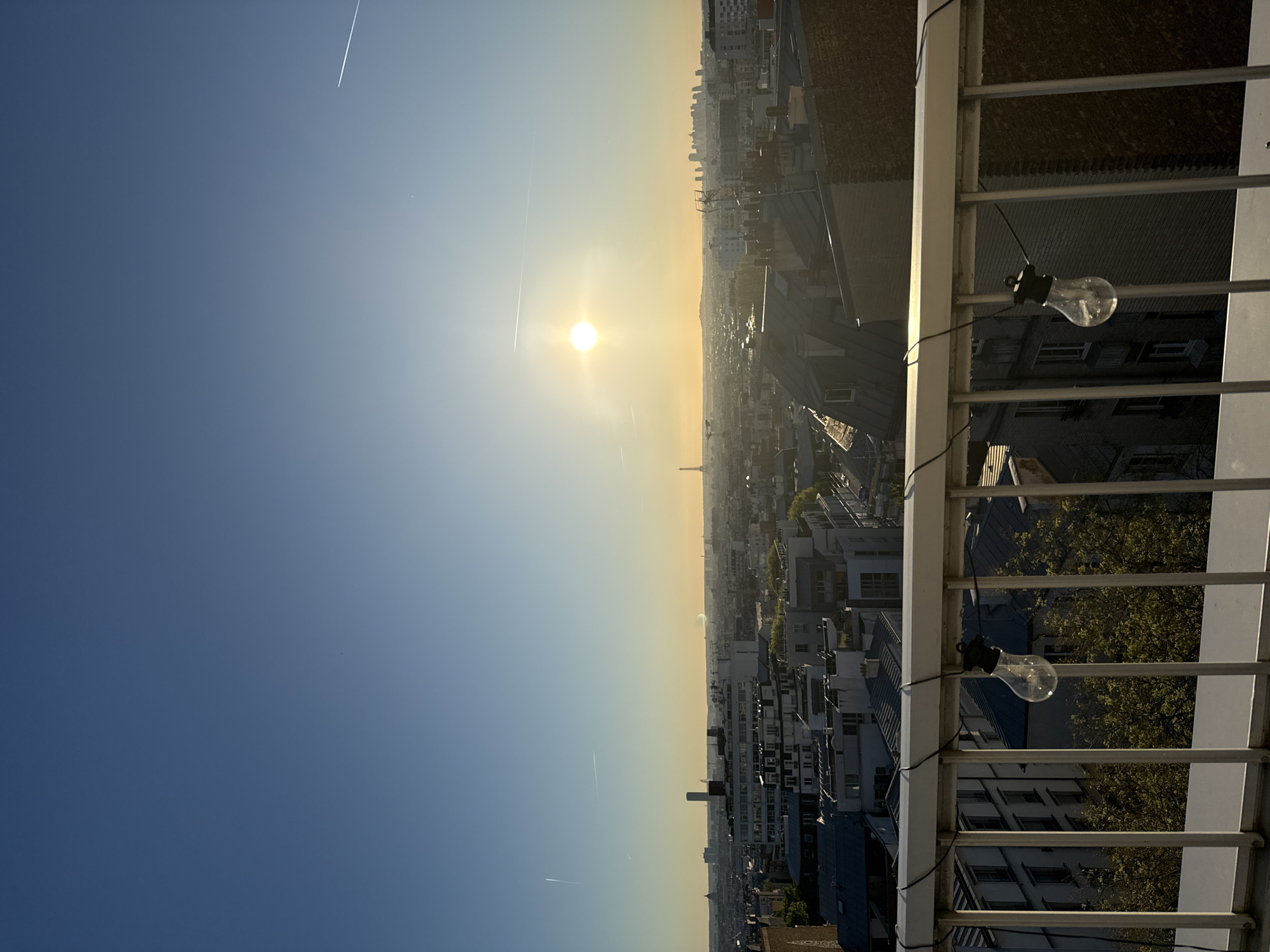 The image depicts a sunrise view from a balcony in Paris, France. The Eiffel Tower is visible on the horizon, appearing as a small dark spire in the golden haze of the rising sun. The city sprawls beneath, a dense collection of buildings with varying heights and roof styles. A few trees are interspersed among the buildings. Two light bulbs hang from the balcony railing in the foreground. The sky transitions from a deep blue to a yellow-orange glow around the sun. A contrail from an airplane is visible in the upper left of the sky.