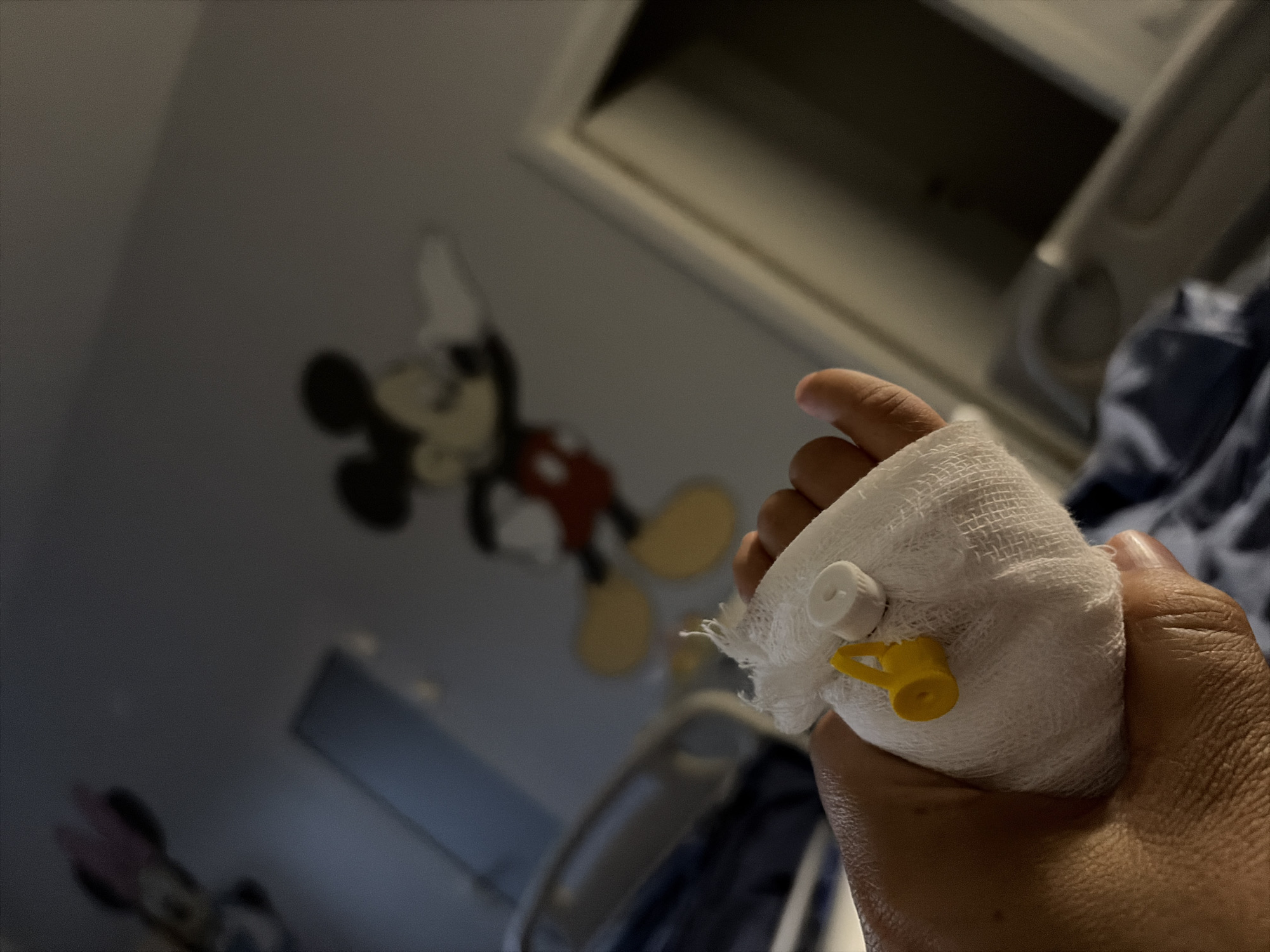 In a dimly lit room, a hand is bandaged with gauze and a yellow port is visible. The background shows a wall with a faded mural of Mickey Mouse and Minnie Mouse. A hospital bed rail is visible on the left. The scene appears to be in a hospital room.