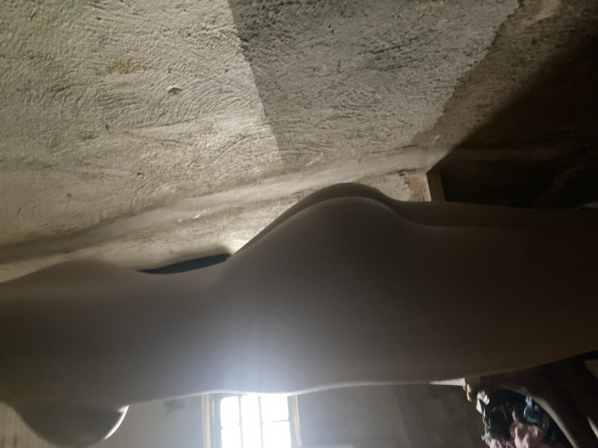 The image shows the side view of a light-skinned mannequin in an indoor setting. The mannequin is positioned to the left, with its back and buttocks prominently visible. The background consists of a rough, textured wall with visible imperfections and a window with a grid pattern. Light streams in from the window, casting shadows on the wall and the mannequin. In the lower left corner, a pile of colorful clothing is visible in what appears to be a basket or container. The overall impression is of a dimly lit, somewhat rustic or unfinished indoor space. There is no indication of Jauro Yinu, Nigeria, in the image.