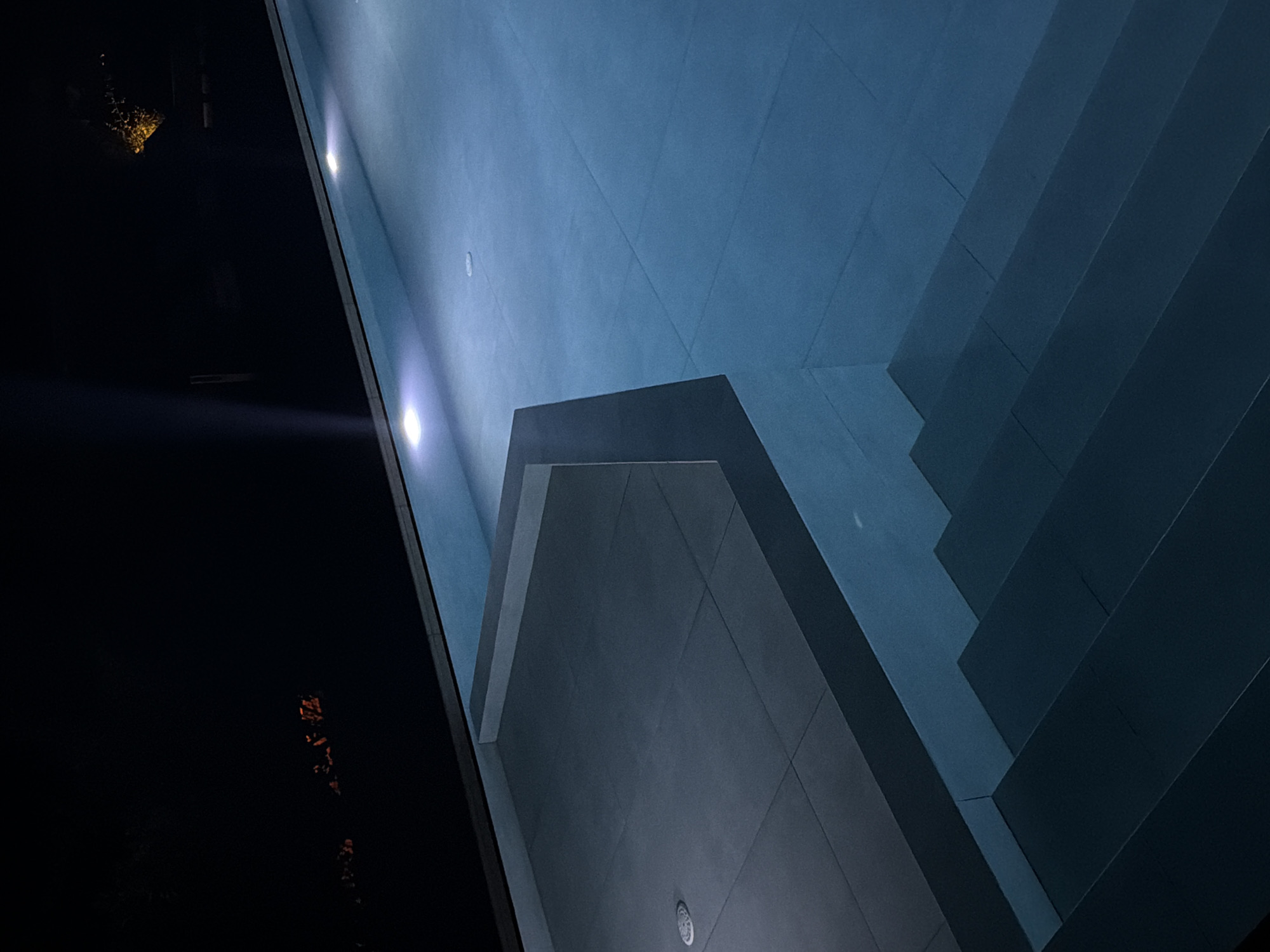 The image shows a modern swimming pool area at night. The pool water is illuminated by soft, white underwater lights, giving it a tranquil blue hue. To the left, there is a tiled, elevated deck area, also lit by subtle lighting fixtures. Steps leading down into the pool are visible in the foreground, appearing dark and well-defined. In the background, the scene transitions into darkness, with faint hints of distant lights, possibly from the town of Viglafia, Greece, suggesting the location is near or within this settlement. The overall impression is one of peaceful, sophisticated nighttime illumination in an outdoor setting.