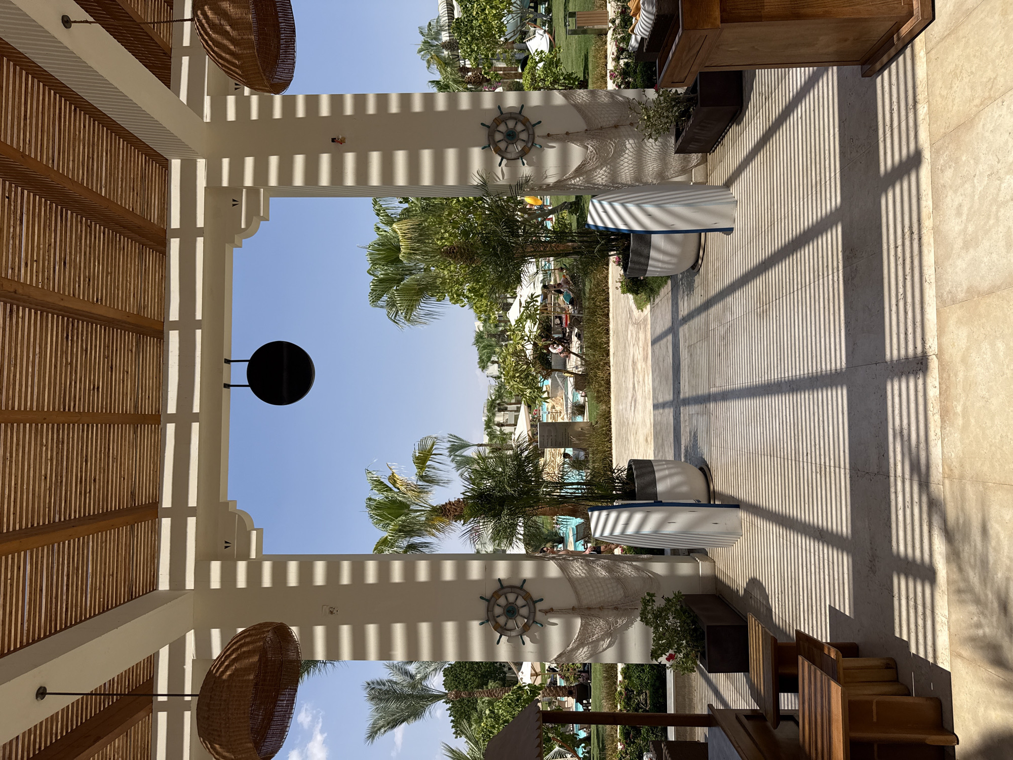 The image depicts an outdoor walkway covered by a slatted wooden roof, casting striped shadows on the tiled floor. The walkway leads towards a resort area with palm trees, lounge chairs, umbrellas, and a swimming pool visible in the background under a clear blue sky. Decorative elements like ship wheels and fishing nets are attached to white pillars. Two tall, white planters with narrow surfboards leaning against them are placed symmetrically on either side of the walkway. The scene suggests a tropical resort environment. The resort appears to be the Hilton Sharks Bay Resort, located in Sharm El Sheikh, Egypt.
