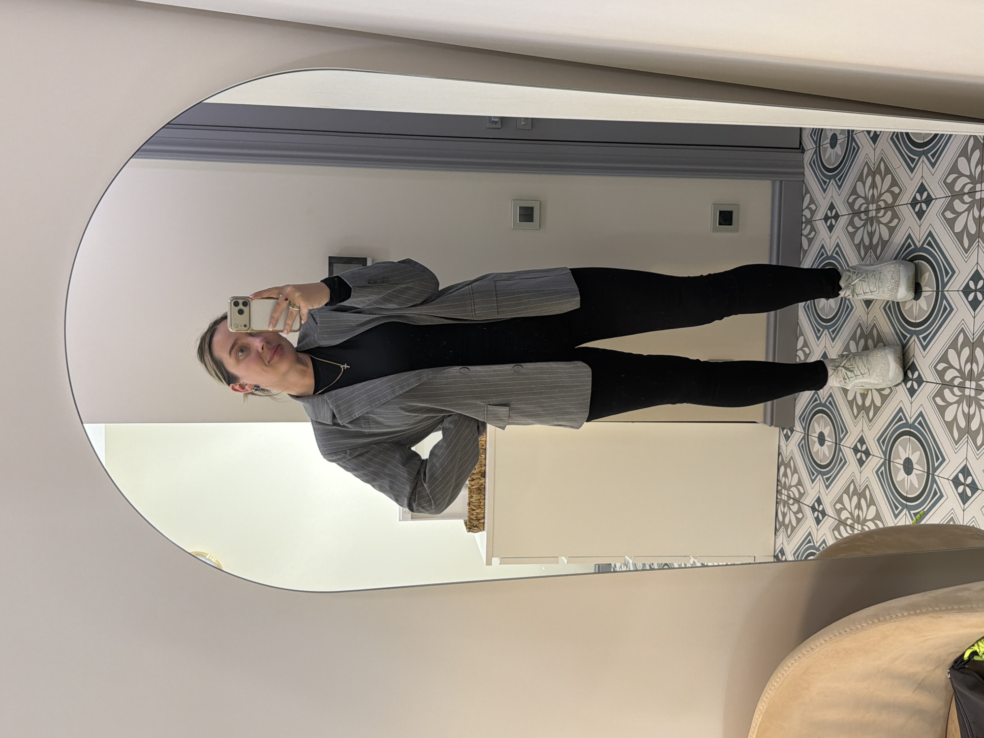 A woman is taking a selfie in a mirror. The mirror is a large, round mirror hanging on a white wall. The woman is wearing a grey pinstripe blazer, black shirt, and black pants. She is also wearing white sneakers. The floor is tiled with a patterned tile. There is a chair and cabinet in the background. Based on the interior and the geometric tile pattern, it could be a bathroom. The image does not contain enough information to determine if the location is in Khimki, Russia.