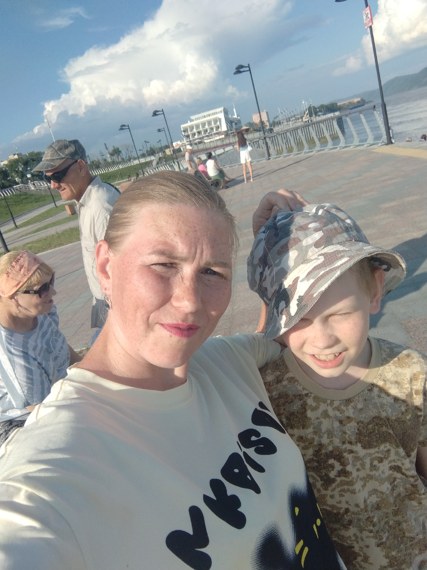 A medium shot shows a group of people on a paved promenade near a body of water. The image is taken from a low angle, with a woman in the foreground holding up a camouflage hat for a young boy. The woman is wearing a light-colored t-shirt with black text. The boy, who appears to be around 8-10 years old, is wearing a camouflage t-shirt and the hat. Behind them, a man in a camouflage hat and sunglasses is visible, along with another woman wearing a headband and sunglasses. In the background, there is a pier, buildings, trees, and a distant hill under a cloudy sky. The overall scene suggests a casual outing on a sunny day.