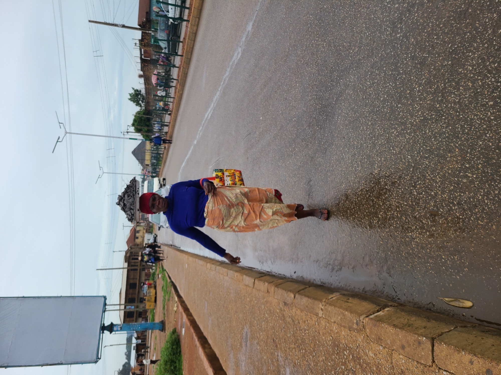 A woman in a blue long-sleeved shirt and patterned skirt is walking along a street in Unknown, Unknown. She has items balanced on her head. The road appears wet. A curb runs along the side of the road, and a blank billboard is visible in the background. In the distance, there are buildings and streetlights.