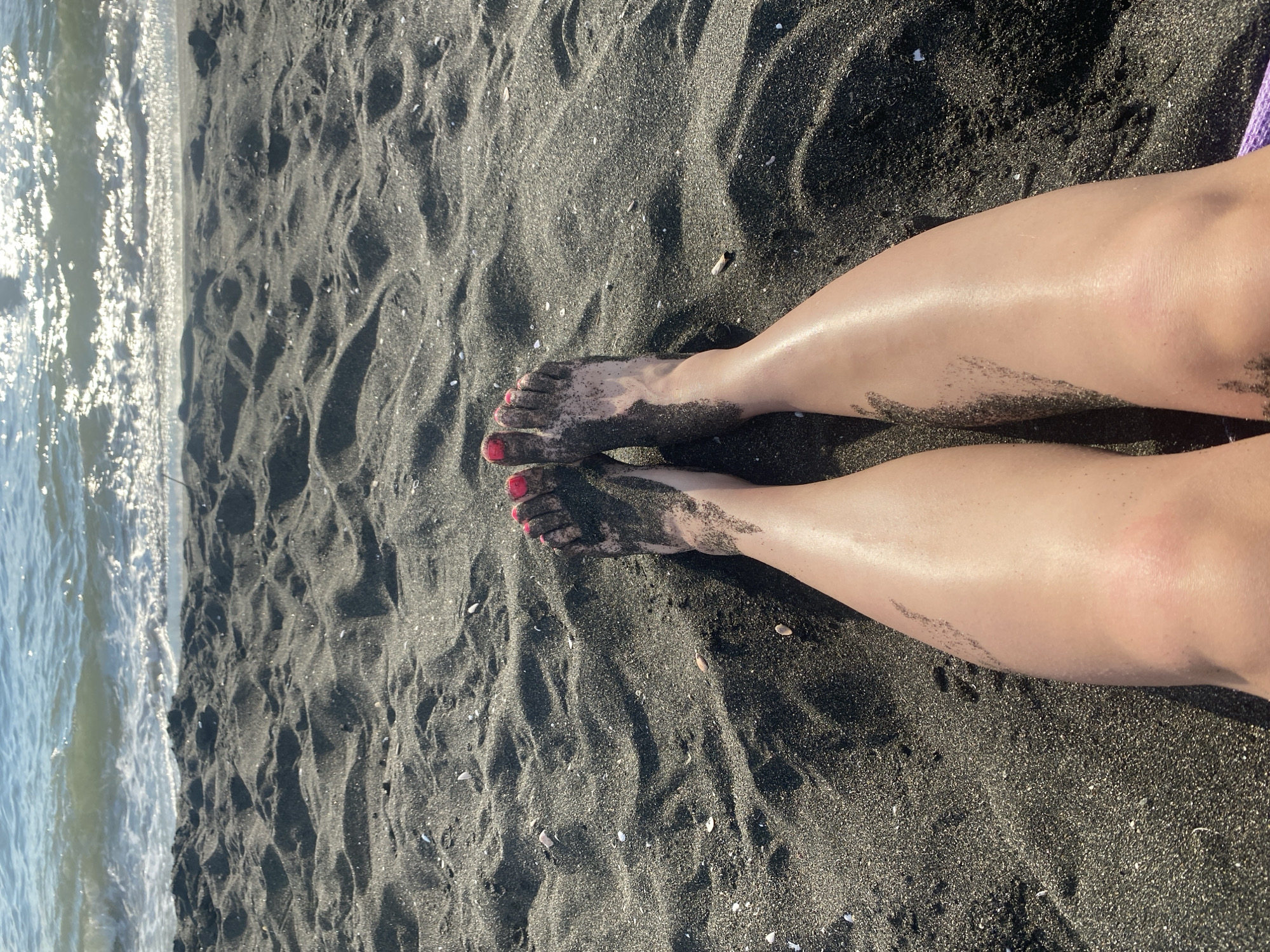 The image shows a pair of bare legs covered in black sand, resting on a beach. The legs are positioned facing the viewer, with the feet closest to the bottom of the frame. The toes are painted with bright pink nail polish. The black sand covers the lower legs and feet, with some lighter patches where the skin is visible. The sand appears to be fine-grained and is rippled, suggesting it has been moved by the wind or water. In the background, the sea is visible, with gentle waves lapping at the shore. The water is a light blue-green color, and sunlight glints off its surface. The overall impression is one of a relaxing day at the beach. The location is the black sand beach of Ureki, Georgia, known for its therapeutic magnetic sand.