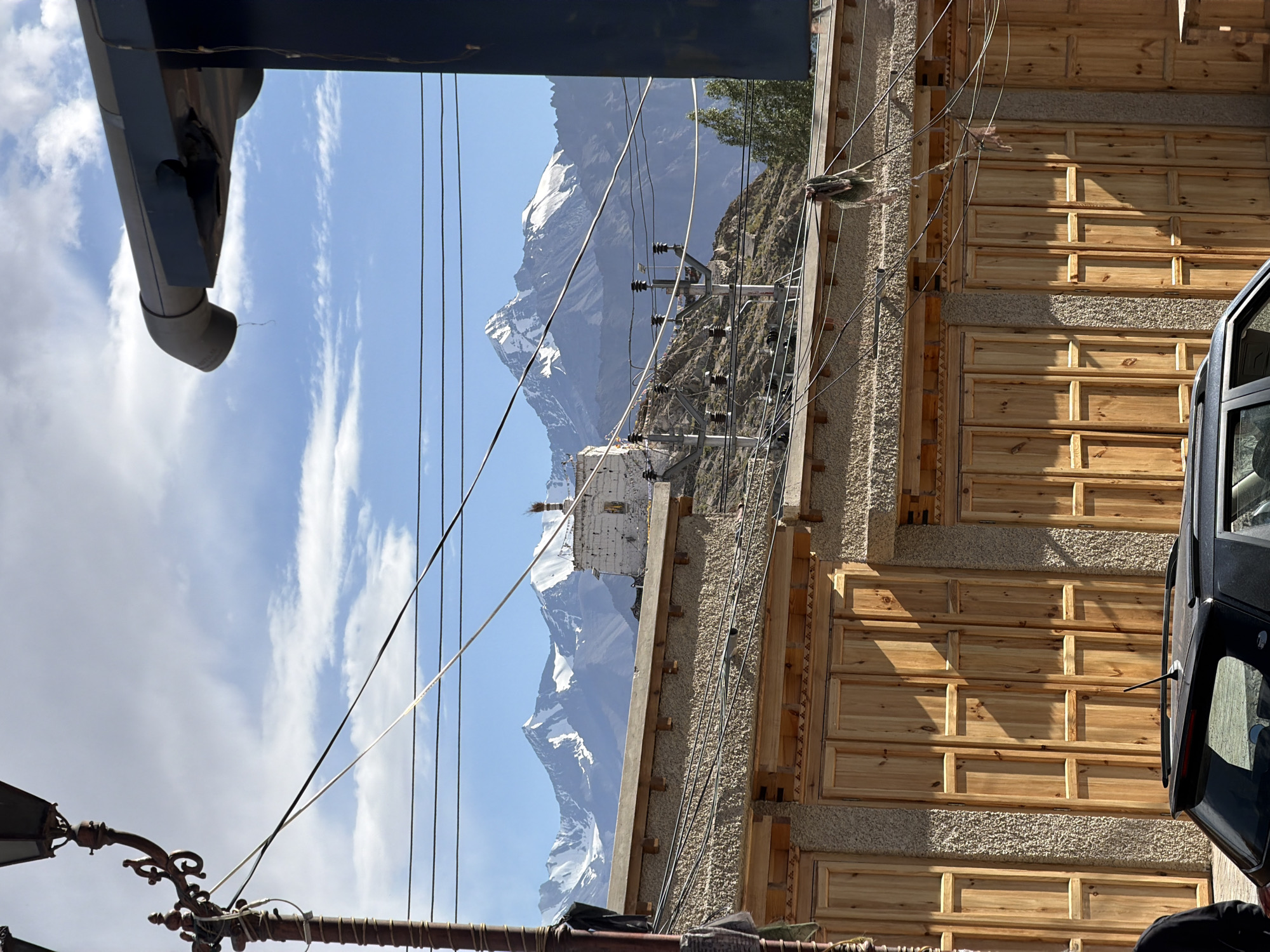 The image captures a street scene in the city of Leh, India, with a focus on traditional Ladakhi architecture and the dramatic Himalayan mountain range in the background. In the foreground, a building with a textured stucco facade and wooden doors is prominently featured. These doors are segmented into vertical panels, showcasing a rustic and natural wood finish. Above the building, a network of electrical wires crisscrosses the frame, indicative of a populated area. A street lamp with ornate detailing stands to the left, adding to the urban element. Further in the distance, nestled against the rugged slopes of the mountains, is a white stupa or monastery, a common sight in the region. The majestic Himalayan peaks, partially covered in snow, dominate the horizon, illuminated by a clear blue sky dotted with white clouds. The overall impression is one of a vibrant street coexisting with the awe-inspiring natural beauty of the Himalayas.