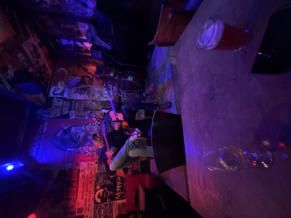 A dimly lit bar with posters covering the walls, two guitarists perform. A drum set sits in the background. A table dominates the foreground, holding a half-full pint glass, a black object, and empty wine glasses. The scene is illuminated with blue and red lights, creating a vibrant yet moody atmosphere. The picture is rotated counterclockwise.