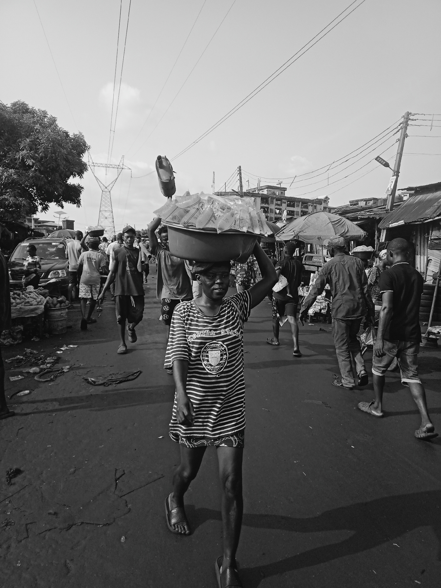 The image depicts a bustling street scene in Onitsha, Nigeria. The foreground is dominated by a young woman walking towards the viewer. She carries a large bowl filled with plastic-wrapped items on her head. She is wearing a striped shirt and sandals. Behind her, several other people are also carrying loads on their heads, navigating the crowded street. There are parked cars and roadside vendors. In the background, buildings and an electrical transmission tower are visible. The road surface appears uneven and littered. The overall atmosphere is that of a busy urban market.