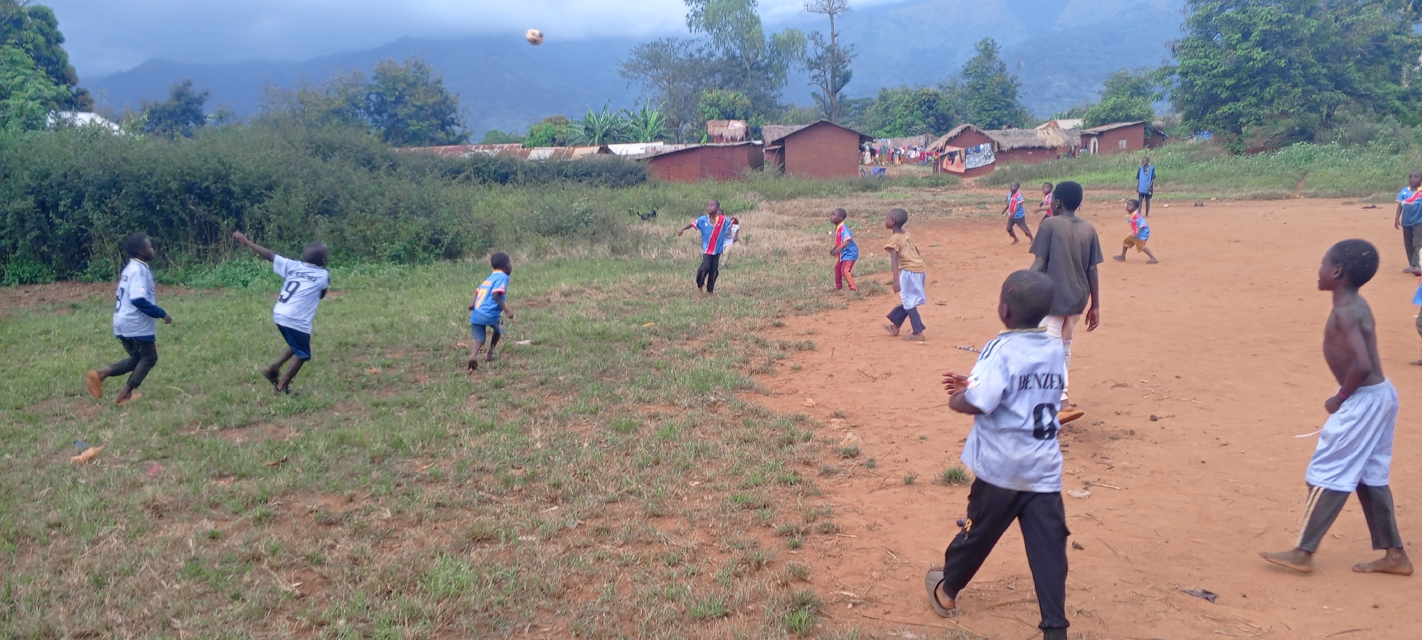 In Ngalula, Democratic Republic of the Congo, a group of approximately 12 children play soccer on a field of mixed dirt and grass. Some children are barefoot or wearing simple sandals, while others have shoes. Some are wearing soccer jerseys; one jersey reads "BENZEMA 8" and another "9". The ball is mid-air above the field. In the background are simple houses with corrugated metal roofs, trees, and mountains in the distance under a cloudy sky. Some people stand in the background between the houses.