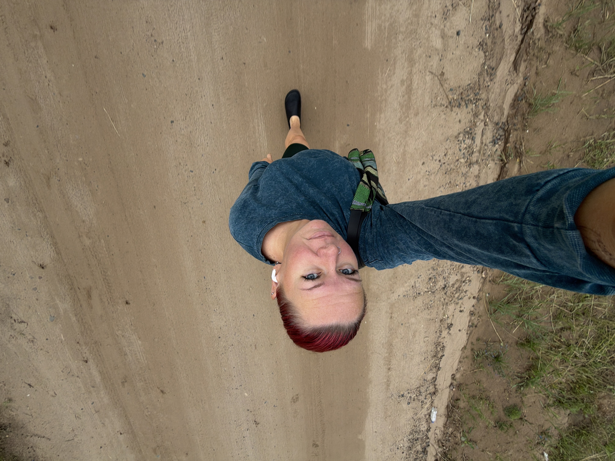 An individual with bright red hair, wearing a dark blue long-sleeved shirt and black shoes, is captured from a high-angle perspective. They are standing on a dirt road, with a patch of grass and soil to their left. The person is looking up towards the camera. A patterned bag is slung across their body. The scene appears to be outdoors, possibly in a rural or less developed area. It is not possible to determine the specific location within Chaikovsky, Russia, from the visual information provided.
