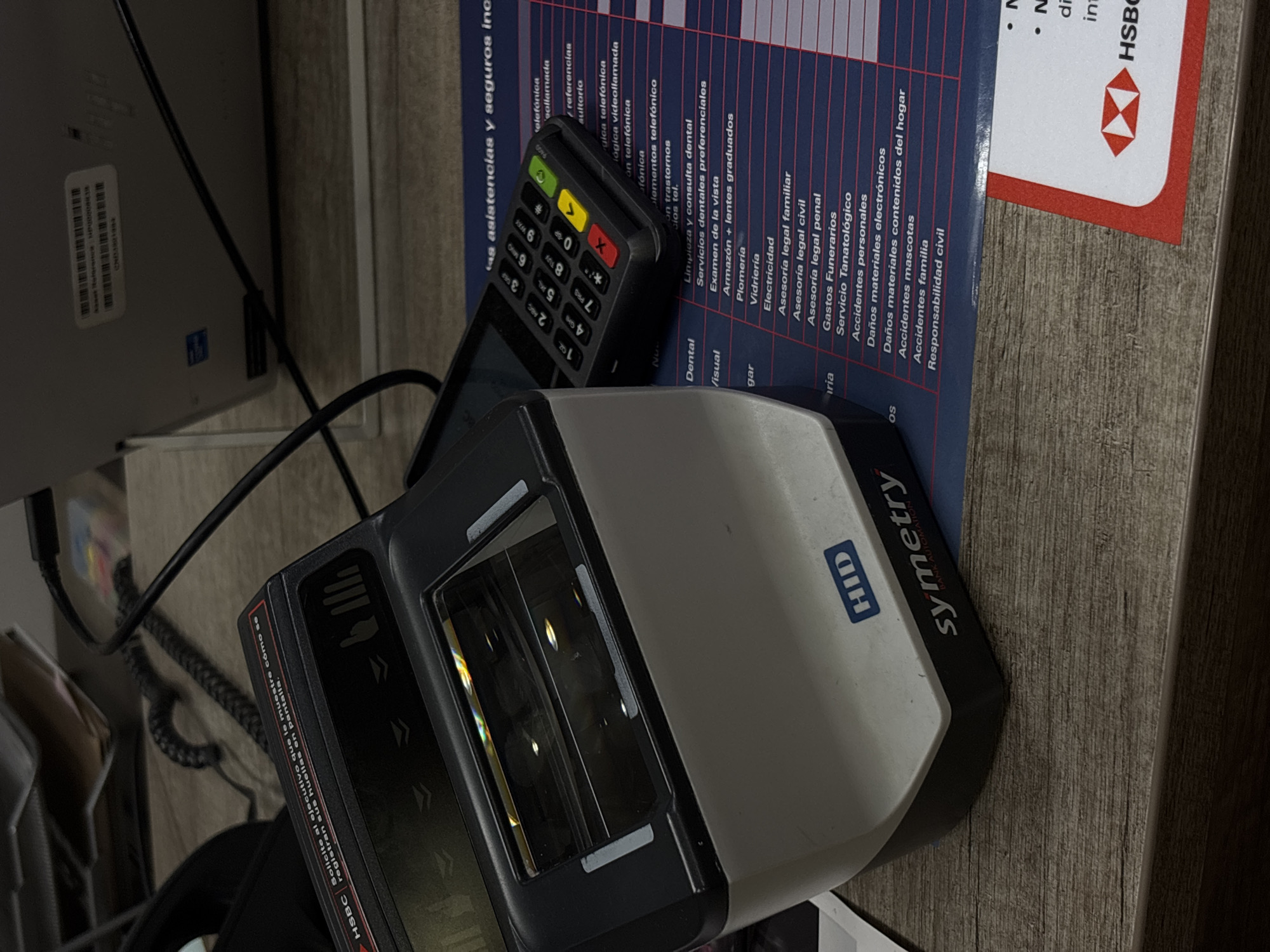 Here is a detailed description of the image:
**Scene Overview:**
The image shows a close-up view of several items placed on a wooden-patterned surface, likely a desk or countertop. The items include a fingerprint scanner, a point-of-sale (POS) terminal, a laptop, and a brochure/booklet. The scene appears to be an indoor commercial setting, possibly a bank or service center, based on the presence of the banking-related items and brochure with HSBC branding. The presence of the spanish text in the image suggests that the location is in a spanish speaking country. The presence of HSBC suggests it is likely located in Mexico City, Mexico.
**Specific Objects and Subjects:**
* **Fingerprint Scanner:** An HID Symmetry fingerprint scanner dominates the foreground. Its biometric scanning window is visible, along with labels and logos.
* **POS Terminal:** A Verifone or similar brand POS terminal is positioned behind the fingerprint scanner. Its keypad and screen are visible, suggesting it is used for payment processing.
* **Laptop:** A silver-gray laptop is partially visible in the background, with a few ports and cable attachments.
* **Brochure/Booklet:** A brochure with a blue color scheme and red HSBC logo is positioned behind the devices. Text in Spanish is visible, detailing insurance-related topics.
* **Cables:** Black cables connect the electronic devices, indicating their power and/or data connections.
**Actions and Interactions:**
It's difficult to infer specific actions from a static image. It could be inferred that a client is currently in the process of completing a transaction requiring biometric identification. The presence of brochures suggests an effort to inform customers about services and policies.
**Environment:**
* **Surface:** The objects rest on a flat surface with a woodgrain pattern, suggesting it is a desk or counter.
**Overall Impression:**
The overall impression is a workspace or point of service at a financial institution, where technology is used to process transactions and manage customer identification, potentially in Mexico City, Mexico.