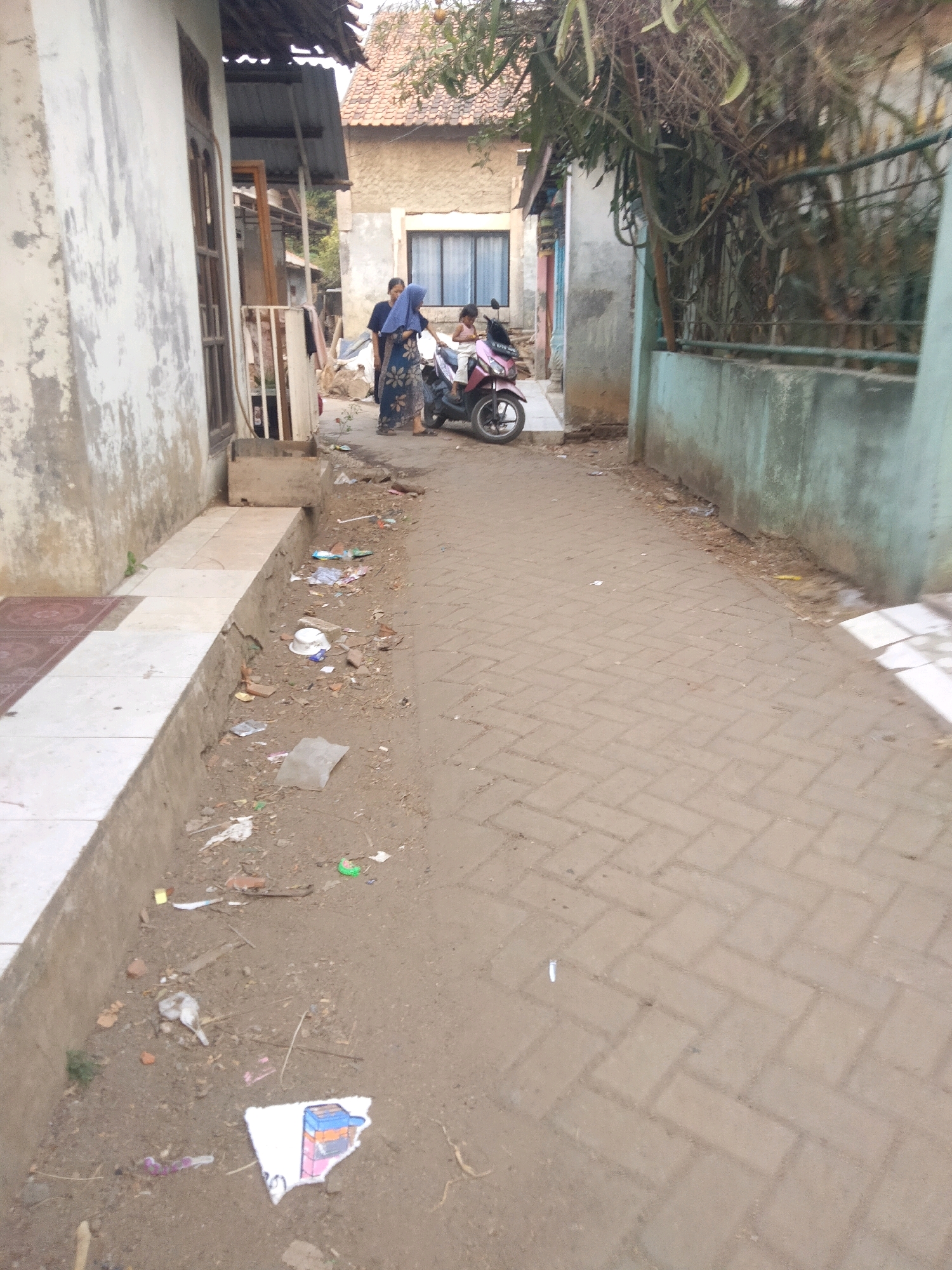 The image shows a narrow alleyway in the Suka Sari area of Indonesia. The ground is a mixture of dirt and brick paving. To the left, there is a light-colored building with a visible drainage channel running along its base. The channel is filled with debris. Further down the alley, two people are standing next to a pink and black motorcycle. One person is wearing a blue hijab and a patterned dress, and the other appears to be a child. They are positioned in front of a tan-colored building with a window. To the right of the alley, there is a light blue concrete wall topped with a metal fence. Vegetation is visible over the fence, and some tree branches hang over the alleyway. The alley is littered with various pieces of trash, including plastic bags, wrappers, and broken items.