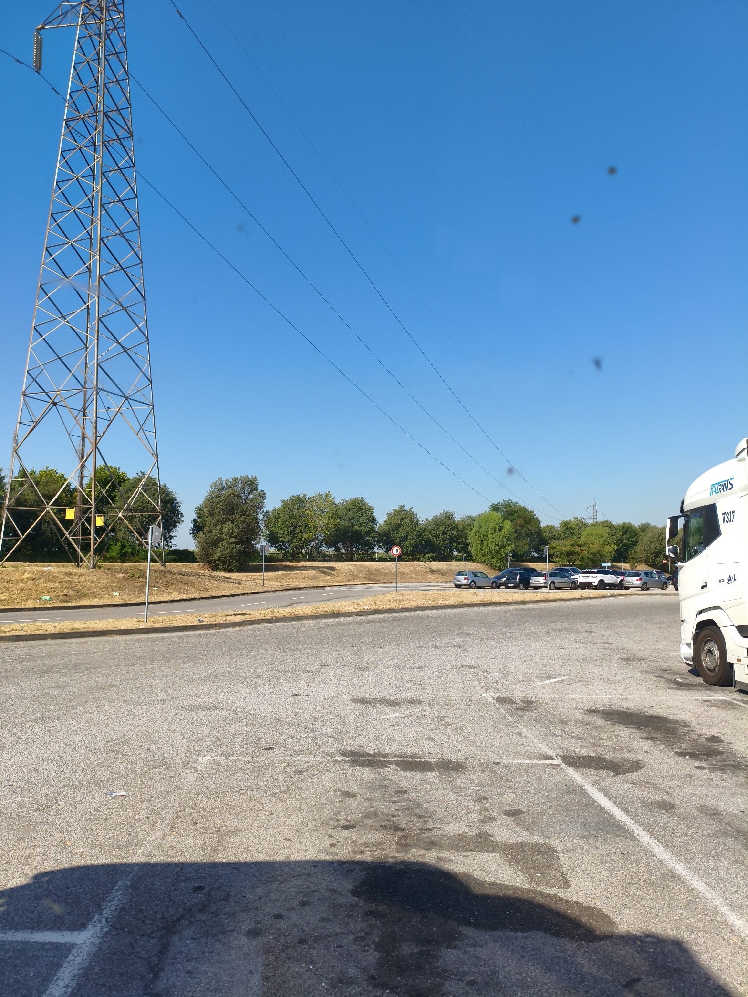 A photograph taken on a sunny day shows a parking lot in Chiari, Italy, with a large metal pylon in the background and a white truck in the foreground. The pylon, a lattice structure of metal beams, stands tall against a clear blue sky, with several power lines extending from it. The parking lot is paved with asphalt and marked with white lines, indicating parking spaces. In the middle ground, a line of cars is parked. The environment is mostly dry, with some patches of grass and trees visible. The overall scene depicts an outdoor commercial or industrial area.