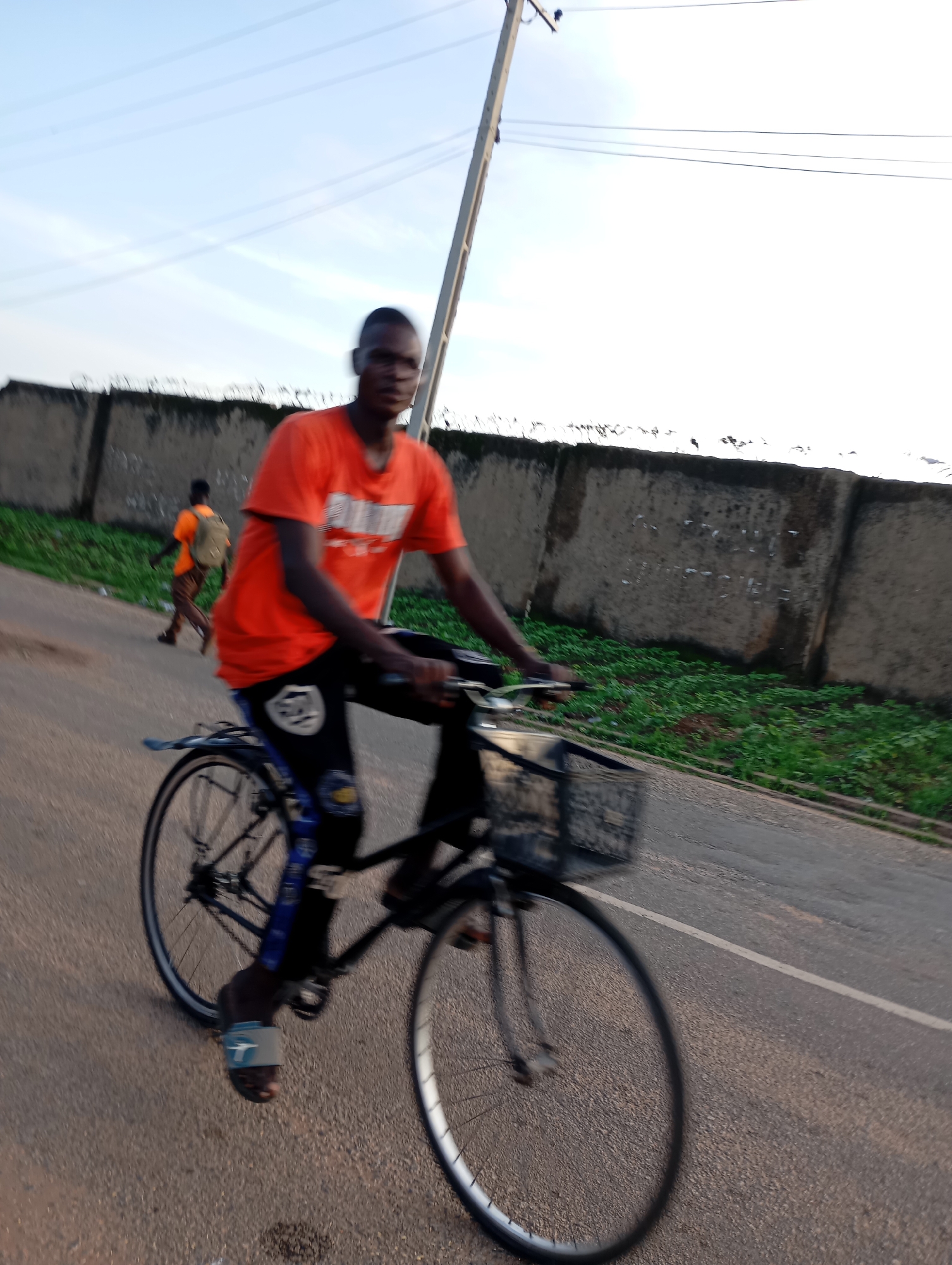 A young man, likely in his late teens or early twenties, is riding a bicycle on an asphalt road. He is wearing an orange t-shirt with a logo that is partially visible and black pants with blue and white accents. His sandals are light blue and white. The bicycle has a black frame and a basket attached to the handlebars. In the background, another young man in an orange t-shirt and carrying a backpack is walking away from the camera.
The scene appears to be outdoors during the day, possibly late afternoon given the lighting. The sky is a pale blue with some light clouds. A concrete wall with barbed wire on top runs along the right side of the road, with green vegetation visible behind it. A utility pole with wires is also present. The road has a white line marking the edge. The image is slightly blurred, suggesting motion, likely from the cyclist.
The overall impression is a common scene in a town or village, possibly in Shendam, Nigeria, as indicated by the location context. The activity is the simple act of traveling by bicycle. There are no strong emotional cues visible due to the blur and the nature of the activity. The visible text on the cyclist's shirt is too indistinct to read.