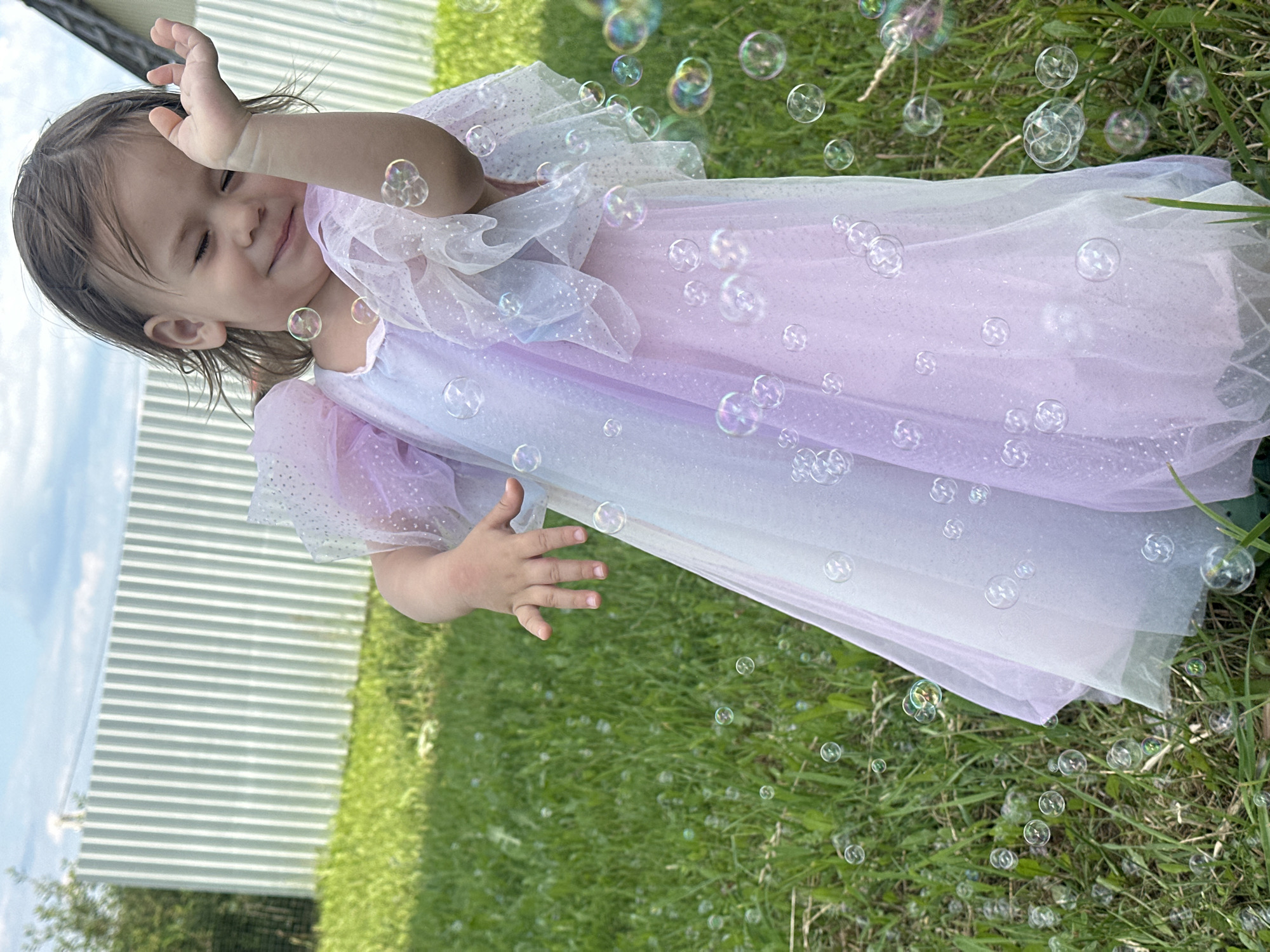 A young child, appearing to be a toddler, is outdoors in a grassy area, possibly a backyard, with a white fence in the background. The child is wearing a shimmering, multi-colored dress with puffy sleeves, predominantly in shades of pink and purple. They are playfully reaching out a hand, with their eyes closed and a smile on their face, as if interacting with a group of bubbles that are floating around them. The ground is covered in green grass, and the sky above is cloudy. The location is identified as Kharnavy, Russia.