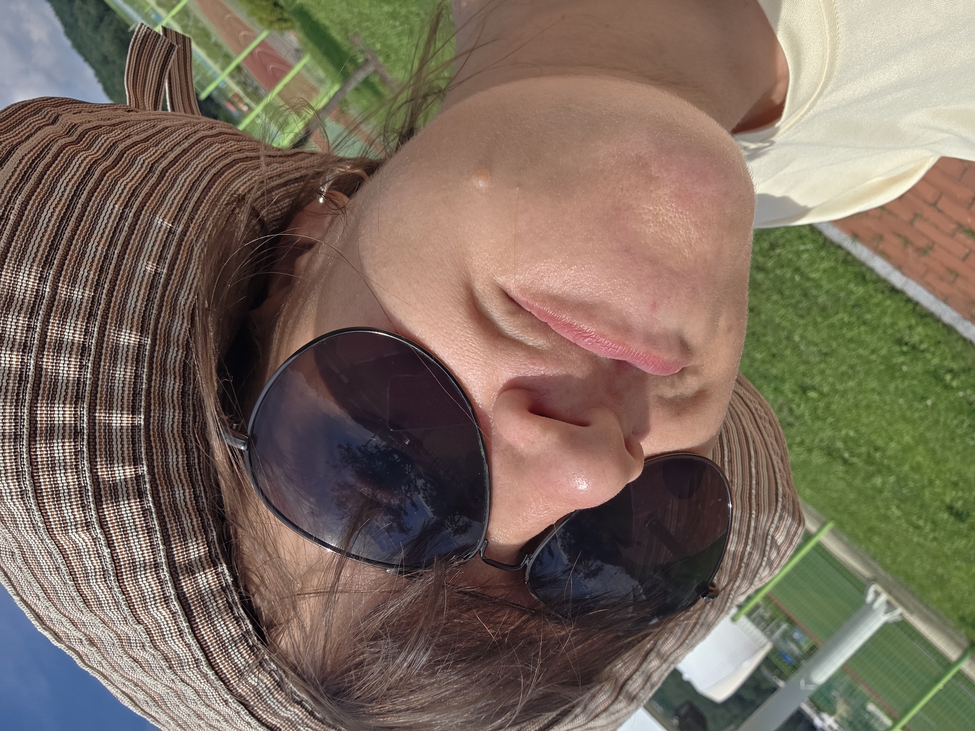 A close-up, low-angle outdoor selfie shows a person wearing a striped brown and white sun hat and dark sunglasses. The person's face is tilted upwards, and their mouth is slightly open. A small, light-colored bump is visible on their cheek. The background consists of green grass, a brick pathway, and a blurred view of a sports field with red and green lanes under a blue sky. The scene appears to be in Dunnae-myeon, South Korea.