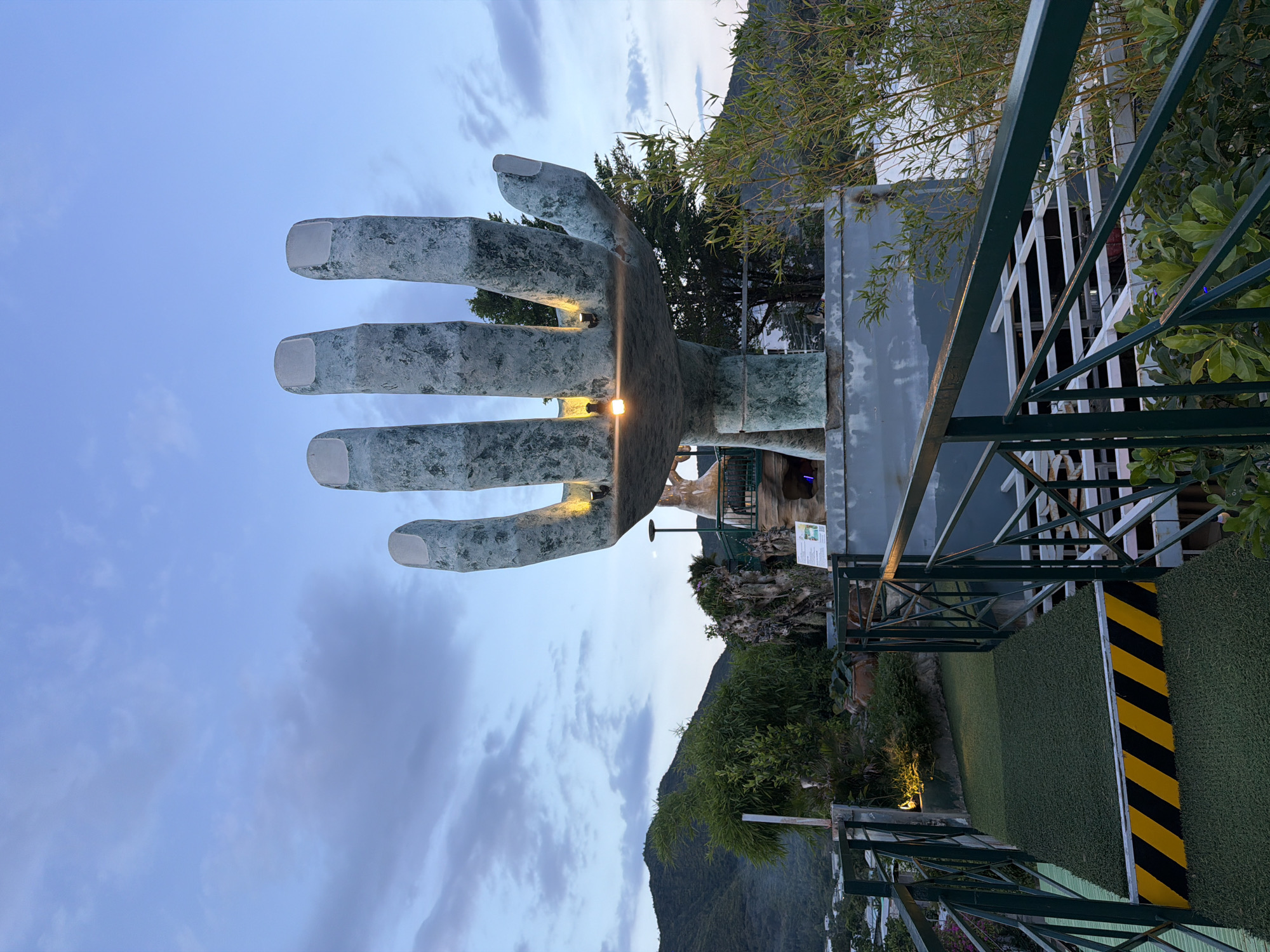 In Nam Nha Trang Ward, Vietnam, at dusk, a colossal concrete hand sculpture, weathered and textured, dominates the frame, its fingers pointing towards a cloudy, pale blue sky. The sculpture, illuminated by small, warm-toned lights embedded in its palm, appears to be situated on an elevated platform or viewing area. A metal railing, painted dark green, encloses the platform, with a walkway covered in artificial turf and marked with yellow and black hazard stripes leading into the scene from the lower left. Beyond the sculpture, lush green hills rise in the background, dotted with trees and structures that suggest a town or village. Some bamboo plants and other greenery are visible in the foreground and around the sculpture, adding to the natural, elevated setting. The lighting and cloudy sky indicate the transition from day to night. No people are visible in the image.