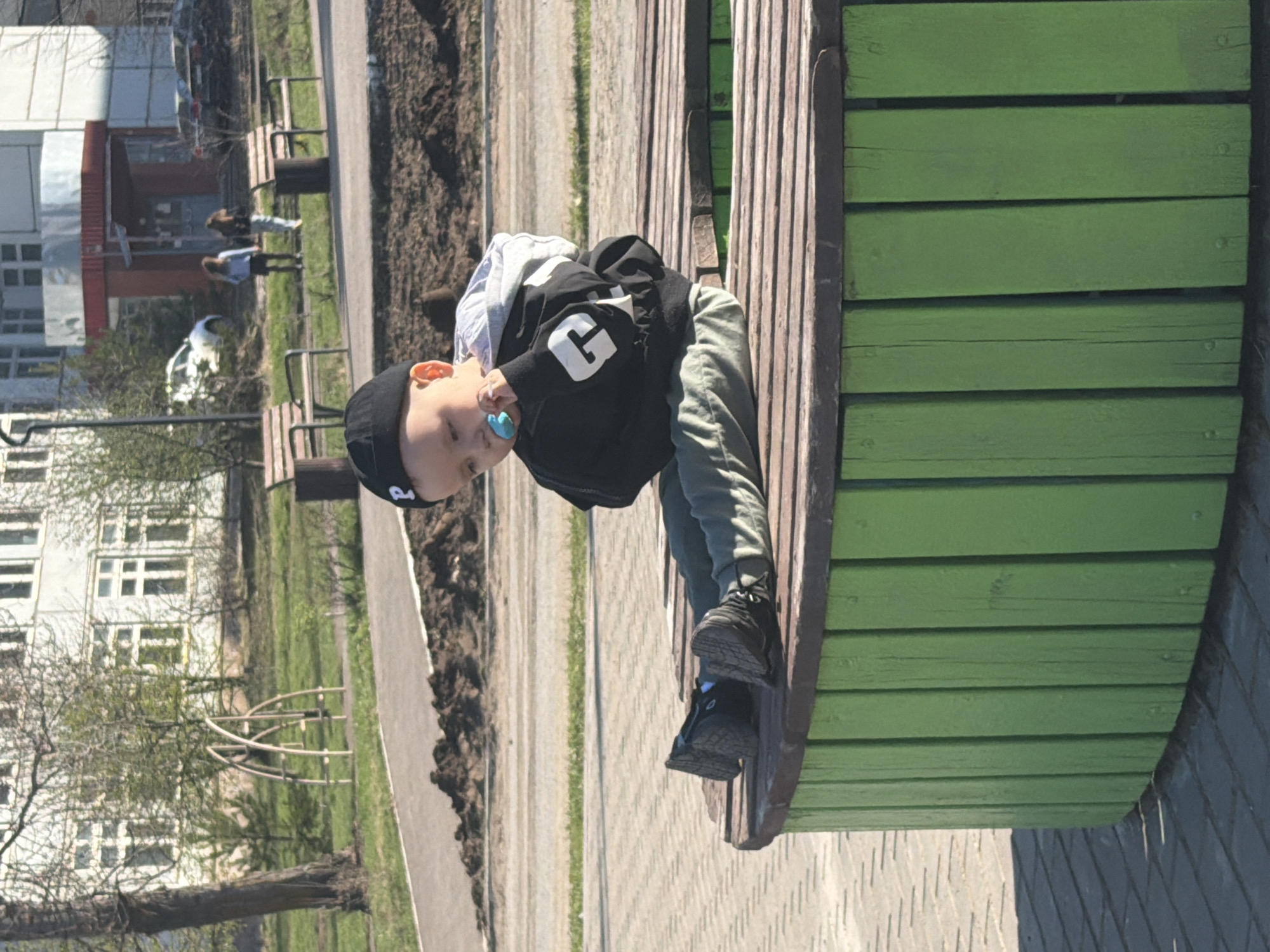 A photograph depicts a young child positioned on a green wooden bench, seemingly located in the city of Chaikovsky, Russia, based on the buildings and foliage visible in the background. The child, who is light-skinned with blond hair, is wearing a black cap and a black vest with a white "G" printed on it, along with light gray pants. A blue pacifier is in the child's mouth. The child's feet, clad in black shoes, are resting on the edge of the bench. The background reveals a gray paved walkway alongside the bench, bordered by a patch of dirt and grass. In the distance, a multistory white building with numerous windows is visible, along with trees, grass, and other park amenities, suggesting the scene is set in a park or public green space.