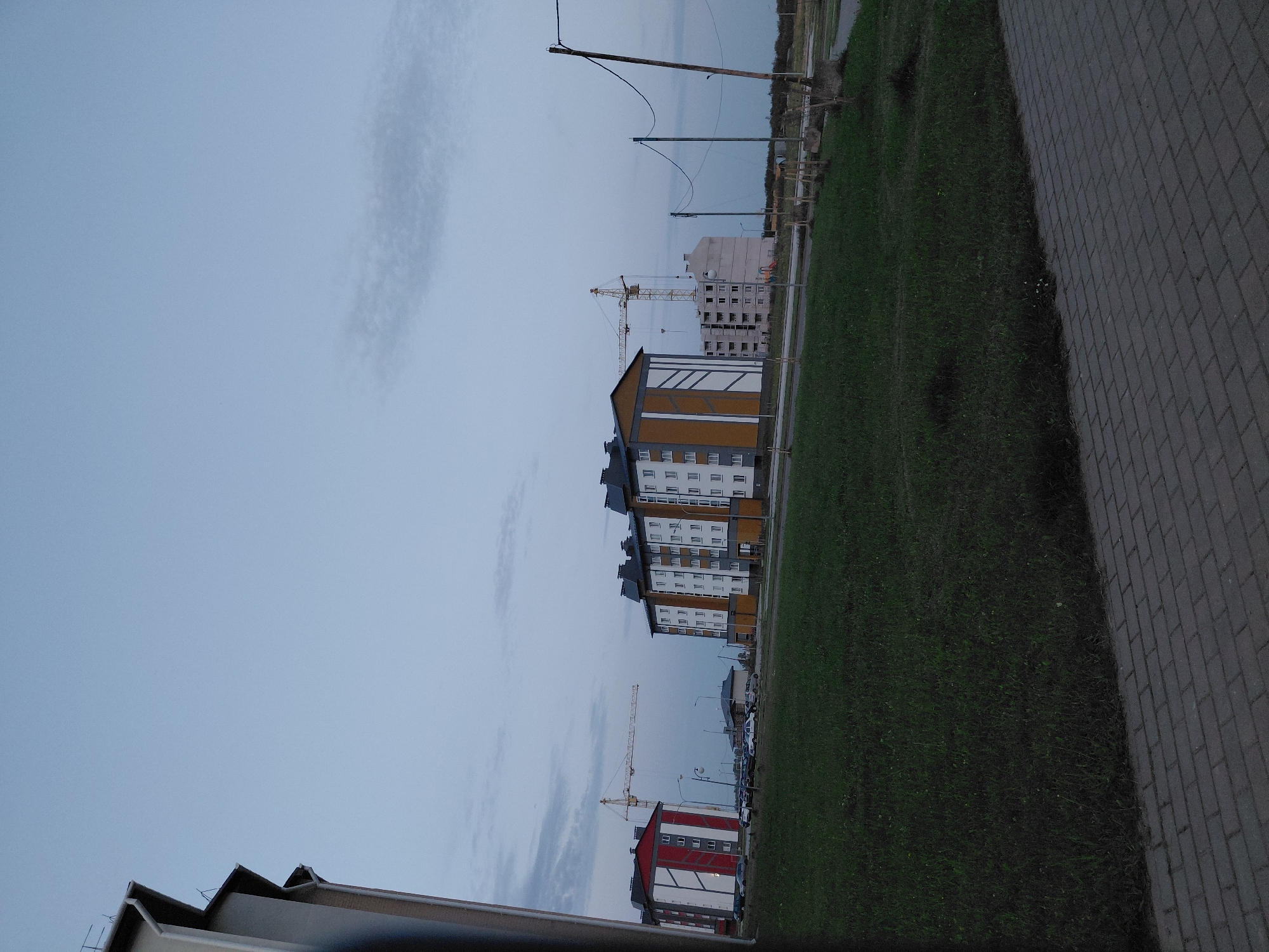 The image depicts an outdoor scene in the city of Smarhon, Belarus, likely during twilight or early evening. In the foreground, a brick-paved walkway runs diagonally from the bottom right corner towards the left. Adjacent to the walkway is a patch of dark green grass.
Dominating the middle ground are several multi-story buildings. On the left, a building with a red and white facade is visible, with cars parked in front of it. To the right of this building, a yellow construction crane extends horizontally across the frame. Further to the right, a larger, lighter-colored building with multiple windows and orange and white accents stands prominently. Another unfinished building with visible window frames is situated behind and to the right of the construction crane. In the background, more buildings and a utility pole with wires can be seen. The sky above is a pale blue with some faint cloud formations. The overall impression is one of a developing urban area.