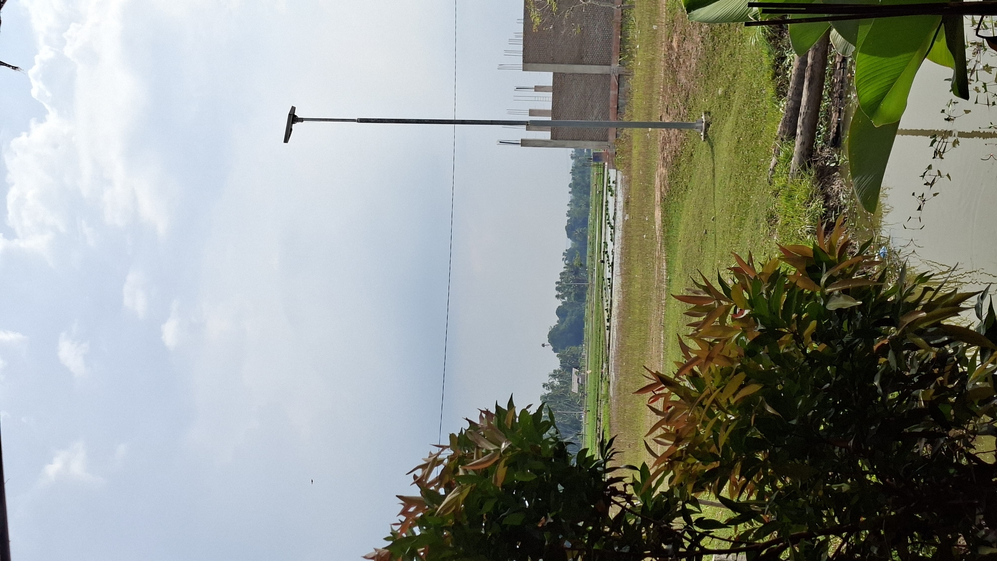 The image depicts a rural landscape under a cloudy sky, likely in Meunasahmee, Indonesia. In the foreground, a lush green bush with reddish-tinged new leaves is on the left. Beyond the bush, a body of water, possibly a canal or flooded field, is visible. In the middle ground, a vast expanse of green rice paddies stretches towards a treeline. A small, simple structure, possibly a hut, is situated within the paddies. A tall, slender lamppost stands in the right-middle ground. To the right of the lamppost, a partially constructed brick building with visible rebar is under construction. In the background, a dense line of trees forms the horizon. The scene suggests a blend of agricultural land and ongoing development.