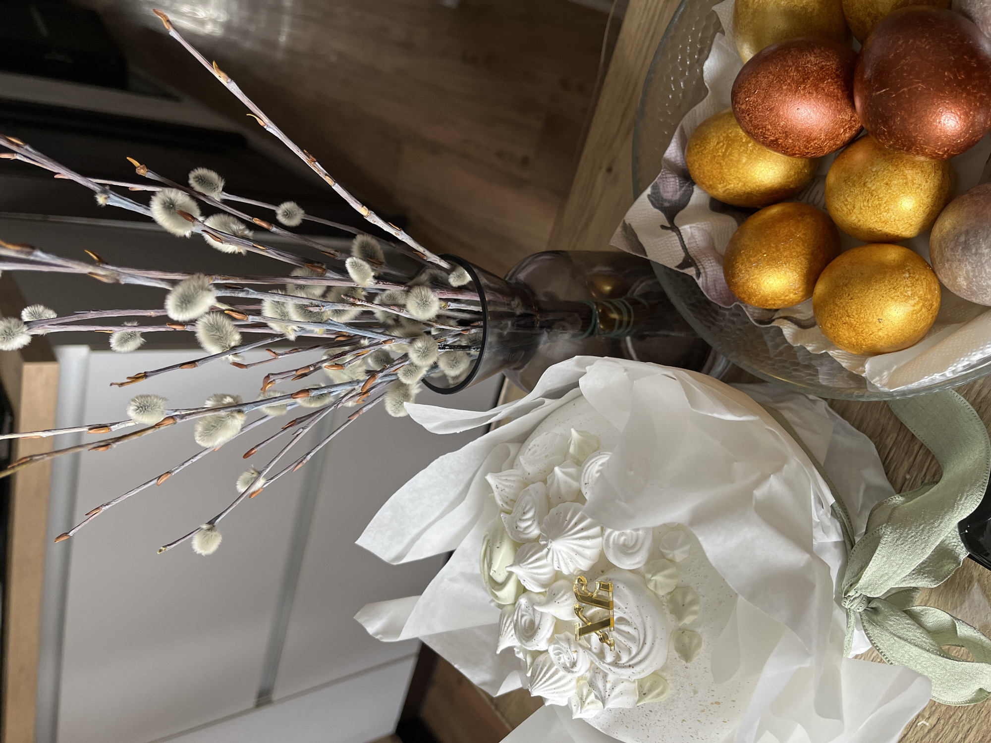 The image shows a still life composition on a light wooden surface. A bouquet of pussy willow branches is arranged in a dark glass bottle. Next to it is a cake decorated with white meringue swirls and wrapped in white tissue paper, tied with a light green ribbon. Adjacent to the cake, a glass bowl contains several Easter eggs with metallic finishes in gold and bronze hues. The setting is likely indoors, possibly a private residence or cafe, but there is no specific information to confirm the location in Krasnoyarsk, Russia.