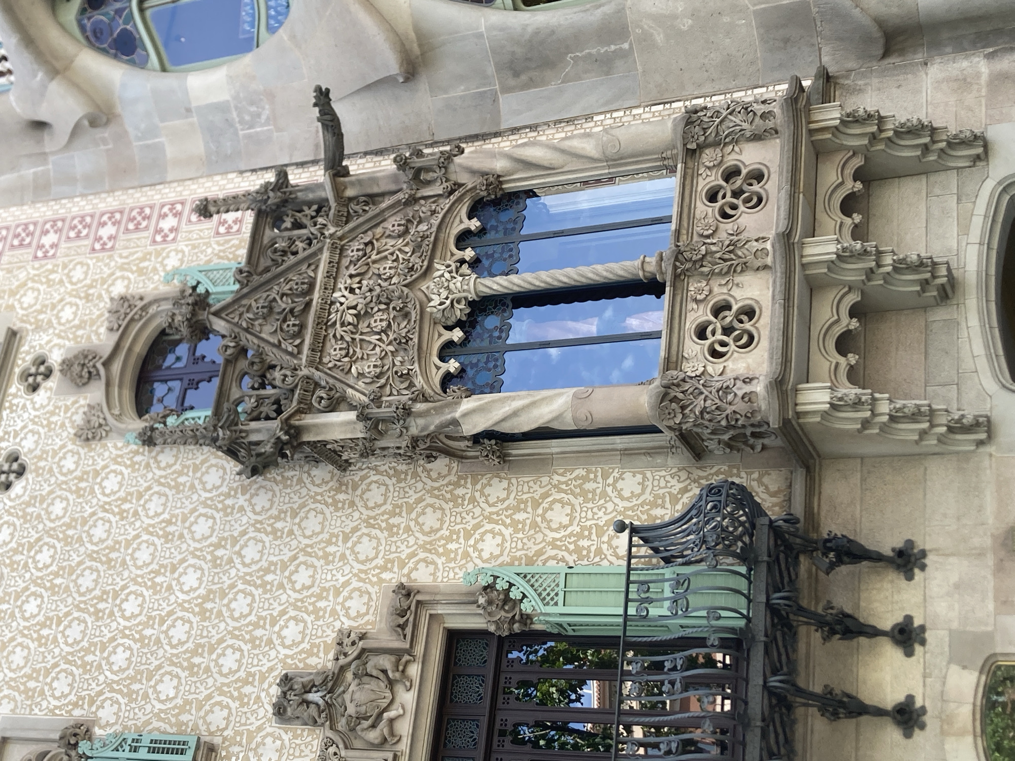 The image shows a detailed view of a building facade in Barcelona, Spain, specifically Casa Batlló, designed by Antoni Gaudí. The architecture is highly ornate, featuring intricate stonework, decorative patterns, and unique sculptural elements.
The main focus is a prominent bay window with a richly decorated stone surround. The window itself is framed by twisted columns and topped with an elaborate, almost gothic-inspired canopy adorned with floral motifs and architectural carvings. A gargoyle-like spout protrudes from the upper right of this structure, suggesting a drainage system. Below the window, a balcony with an iron railing is visible, supported by decorative brackets. To the left of this central element, another window is partially visible, with a dark wooden frame and a green shuttered window next to it, a detail also echoed on the balcony. The building's facade is covered in a patterned stucco or tile work, a beige background with a repeating circular and star-like motif in a lighter shade. Adjacent to this patterned section, a wall of lighter gray stone blocks is visible, with a large, rounded window featuring stained glass in shades of blue.
The lighting suggests it is daytime, with sunlight highlighting the textures and details of the building. The sky reflected in the window is a clear blue, indicating good weather. There are no people visible in the image. The image captures a close-up, architectural perspective, emphasizing the craftsmanship and artistic style of the building.