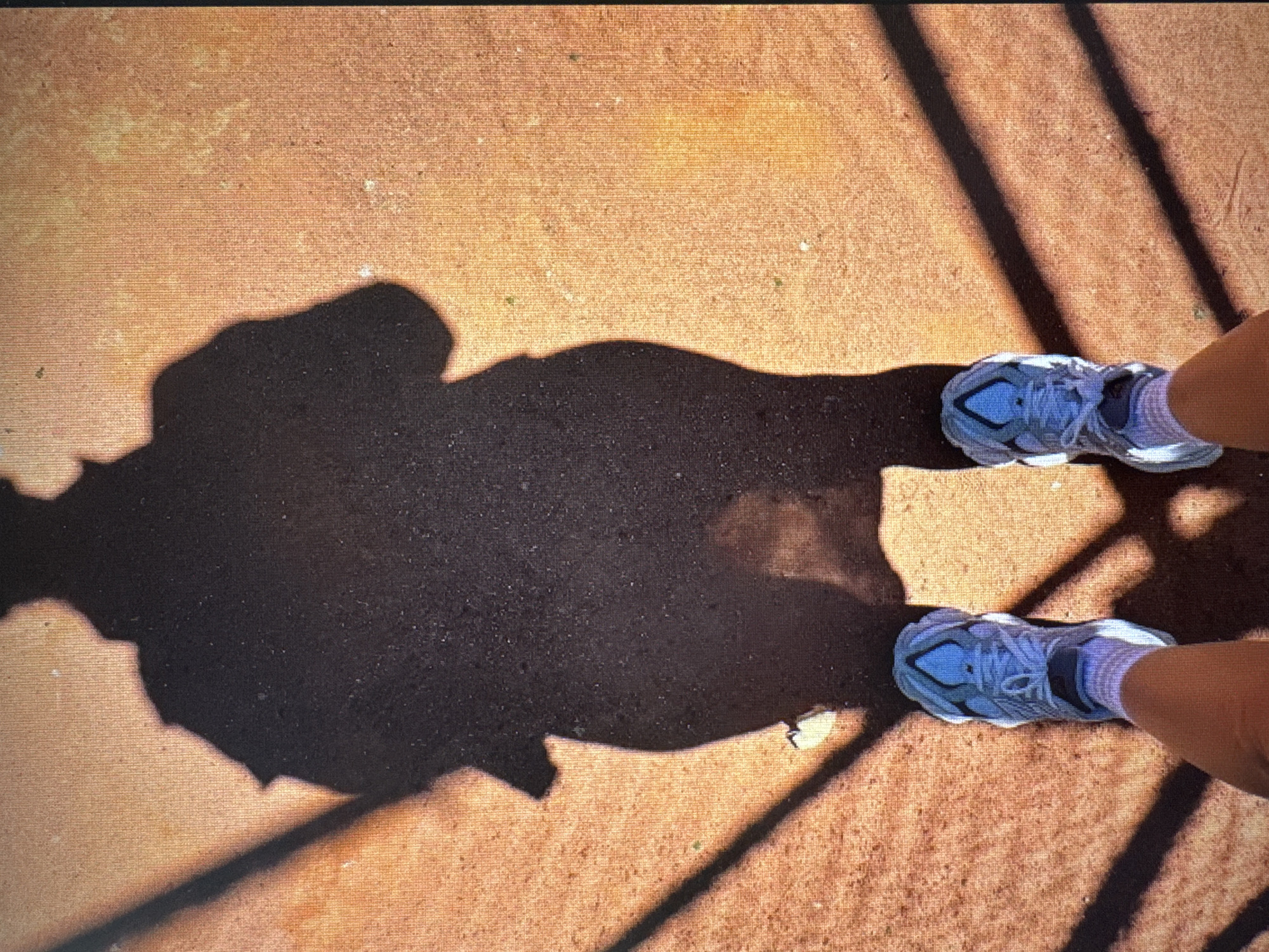 An overhead shot captures a person's feet and their prominent shadow on a textured, reddish-brown ground surface, located in Vratsa, Bulgaria. The individual wears light blue or grey athletic shoes with white soles and white socks, with the lower part of their legs visible above the ankles. A large, dark shadow of a person, likely the photographer's, extends from the left side of the frame across the ground.
Bright sunlight is evident from the sharp, well-defined shadows, indicating a sunny day, likely during mid-morning or early afternoon. Additionally, multiple parallel shadows, suggestive of bars, a fence, or railings, are cast from the upper right corner, diagonally intersecting the ground, the person's feet, and the main shadow. The ground material appears to be a paved or artificially surfaced area, possibly a sports court or pathway. The image portrays a moment of standing, perhaps during a walk or break, with the camera directed downwards. There is no visible text in the media file.