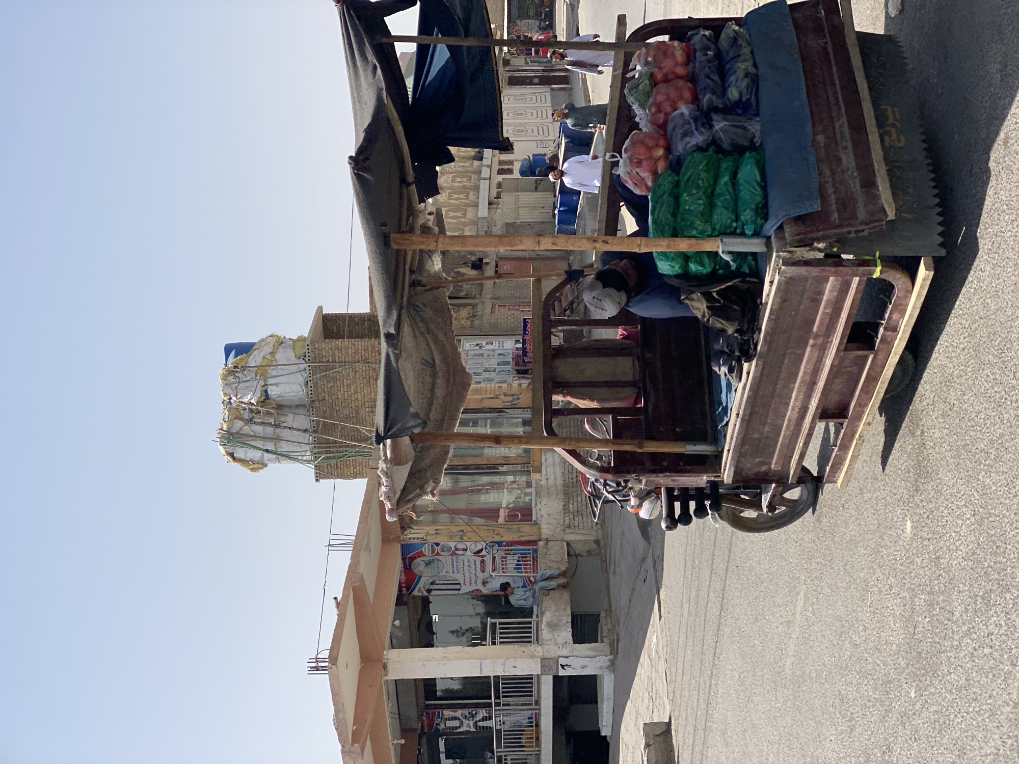The image depicts a street scene in Kandahar, Afghanistan, under a clear, bright sky. In the foreground, a three-wheeled cart is parked on the asphalt road. The cart is laden with produce, including several clear plastic bags filled with red onions and green vegetables. A dark tarp is stretched over a wooden frame to provide shade for the cart's contents and possibly the driver. Partially visible inside the cart is a person wearing a dark blue shirt and a cap, appearing to be resting or attending to the goods. To the left of the cart, a building with a covered walkway and shops is visible. A man in light blue clothing is seated on a ledge outside one of the shops, looking down at something in his lap. Further down the street, several men in traditional Afghan attire are walking. The architecture suggests a typical urban environment in the region, with brick and concrete structures. The overall impression is of a quiet, daytime street in a city in Afghanistan.