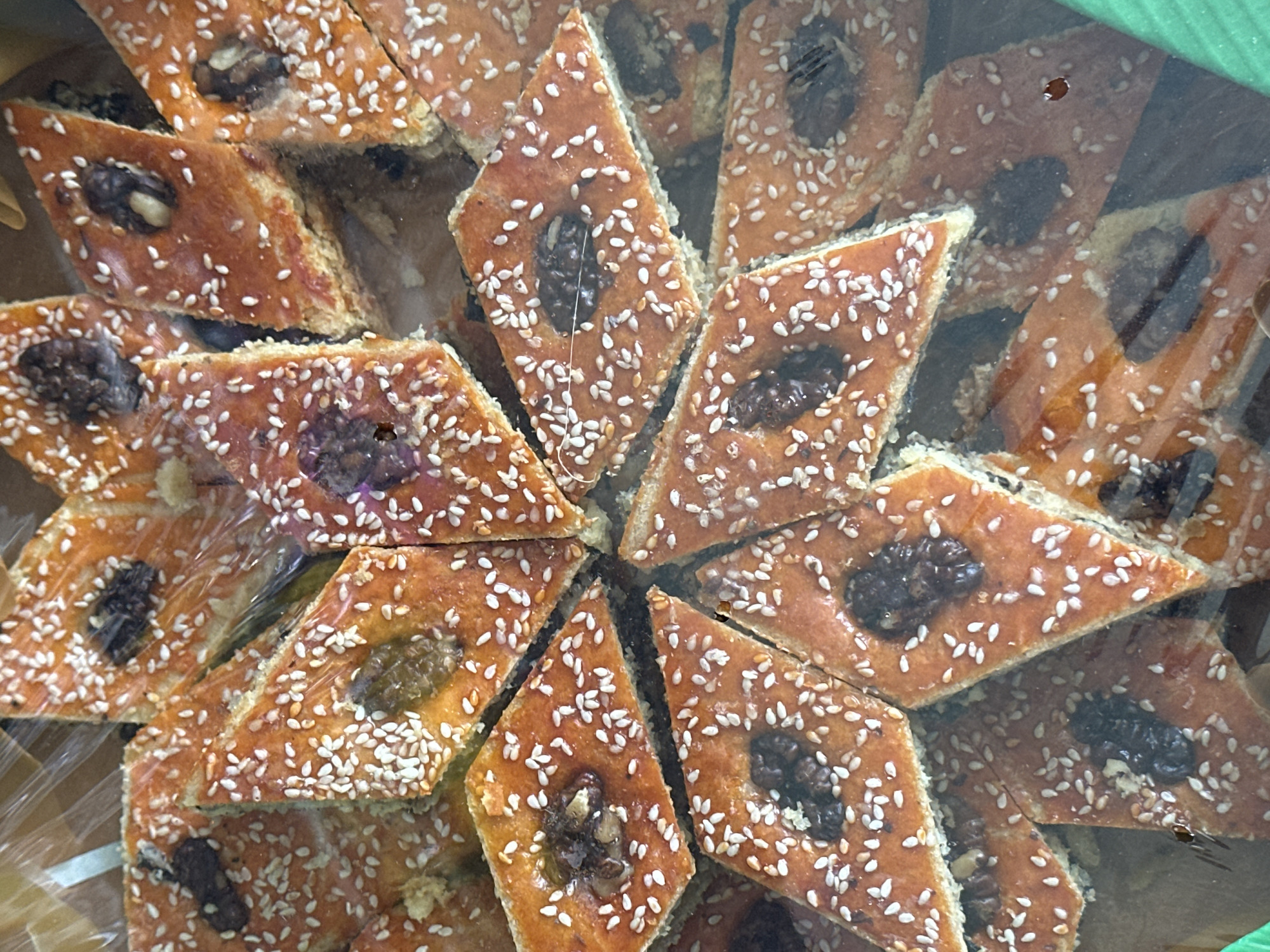 In a close-up, overhead shot, a collection of diamond-shaped pastries, likely baklava or a similar sweet treat, are densely packed in a transparent plastic container. The pastries have a golden-brown, glazed top, sprinkled generously with white sesame seeds. Embedded within each pastry is a dark, textured piece, appearing to be a walnut half. The baked goods are layered and slightly overlapping, creating a visually rich texture. The lighting highlights the sheen of the glaze and the distinct grains of the sesame seeds. The scene appears to be a retail display, possibly in a bakery or market, within the city of Özgön, Kyrgyzstan, though no specific city landmarks are visible. The focus is entirely on the food item, with the surrounding environment obscured by the plastic packaging and shallow depth of field.