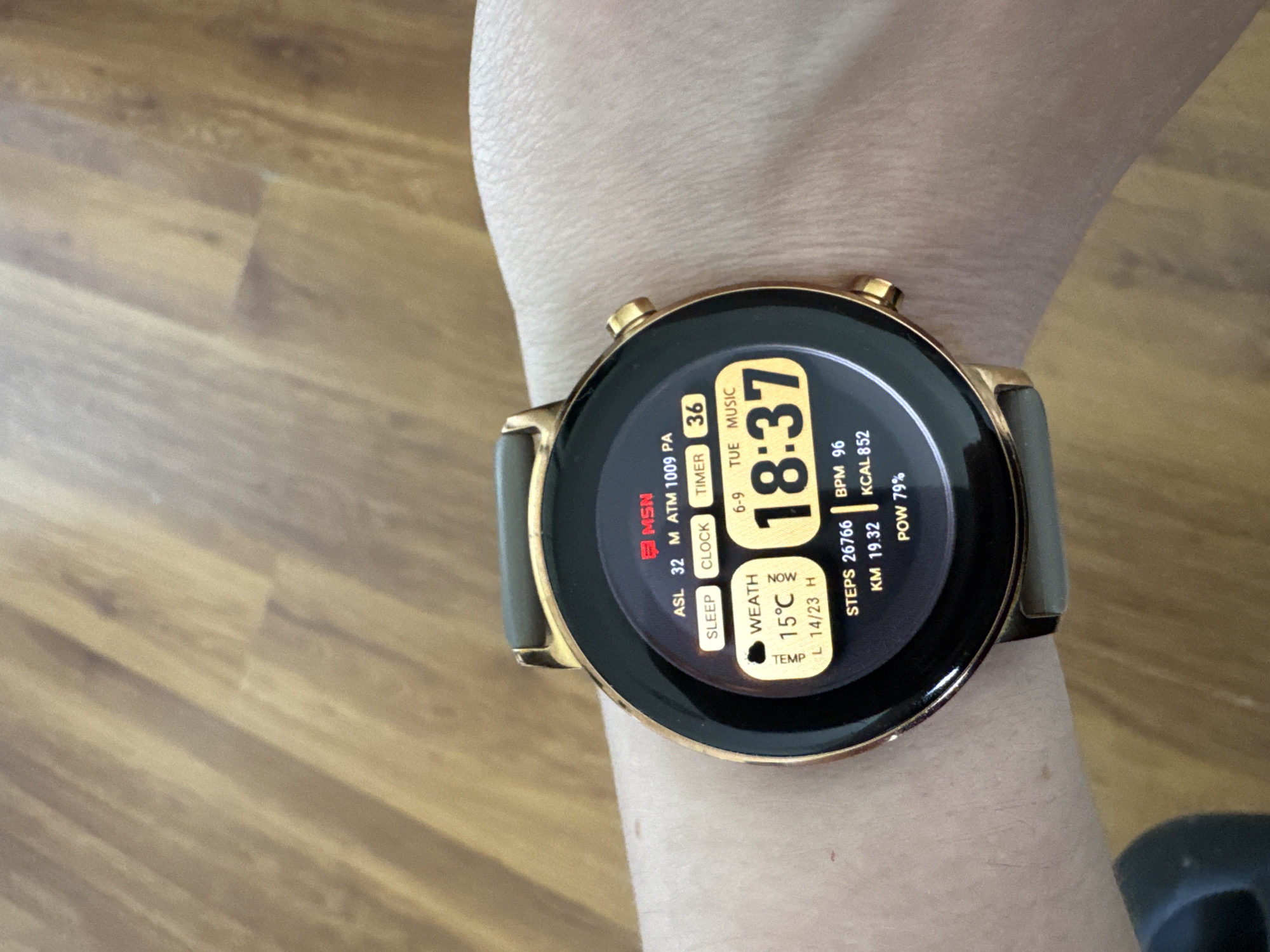 The image shows a close-up view of a person's wrist wearing a smartwatch. The smartwatch has a round face with a gold-colored bezel and a grey strap. The screen displays various information including the time "18:37", steps count "26766", heart rate "BPM 96", and battery level "POW 79%". There are also icons and text for weather, sleep, clock, timer, and music. The background appears to be a wooden surface, possibly a floor or table. The image does not contain any identifiable landmarks or context that would allow for the determination of its location in Riga, Latvia.