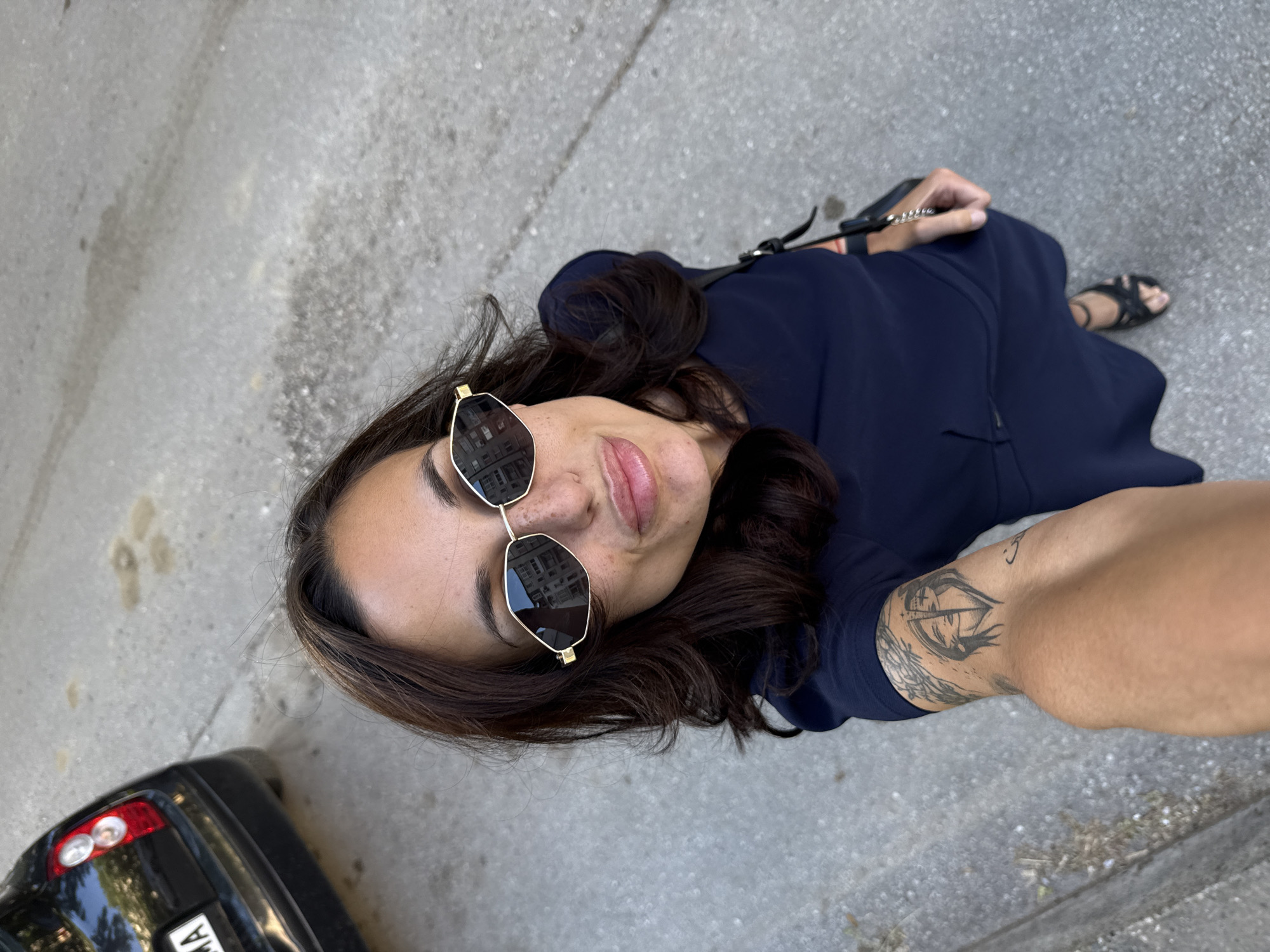 A woman is taking a selfie from a high angle while standing on a sidewalk. She is wearing a dark blue dress and black sandals. Her hair is dark brown and she is wearing sunglasses with gold frames. She is holding a black purse in her hand. A car is parked on the left side of the frame. The background appears to be a sidewalk and a road. The location is Pleven, Bulgaria. There is a tattoo on her right arm.