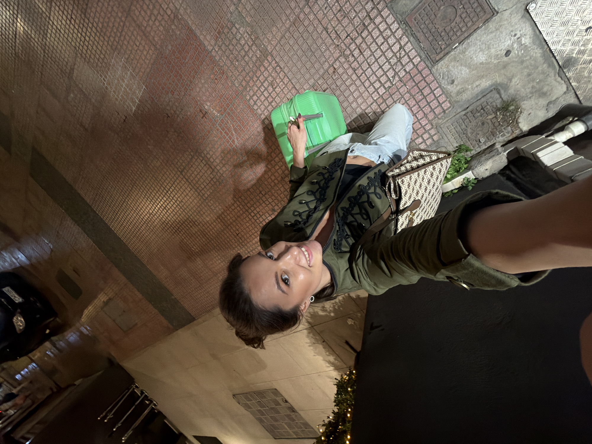 A woman takes a low-angle selfie at night in an urban outdoor setting in Athens, Greece. She is smiling broadly and looking directly up at the camera. Her dark hair is pulled back, and she wears a single silver hoop earring. She is dressed in a dark olive green military-style jacket with elaborate black braided and buttoned trim on the front. Underneath, a light-colored top is visible. She is also wearing light blue pants or jeans.
To her left, she holds a bright lime-green suitcase by its extended handle. A patterned shoulder bag with light brown and beige tones, featuring repeating logos (possibly "Guess"), hangs from her shoulder.
The ground is paved with a mix of reddish-brown square tiles arranged in a grid pattern and sections of grey concrete. Several metal grates and manhole covers are visible on the ground. Small patches of green foliage grow near these covers. In the background, there are dark buildings or structures, and on the far left, part of a dark-colored vehicle is visible. On the right, decorative warm-toned lights, possibly from a Christmas tree or other festive display, add illumination. The overall impression is that of a traveler pausing in a city street or sidewalk.