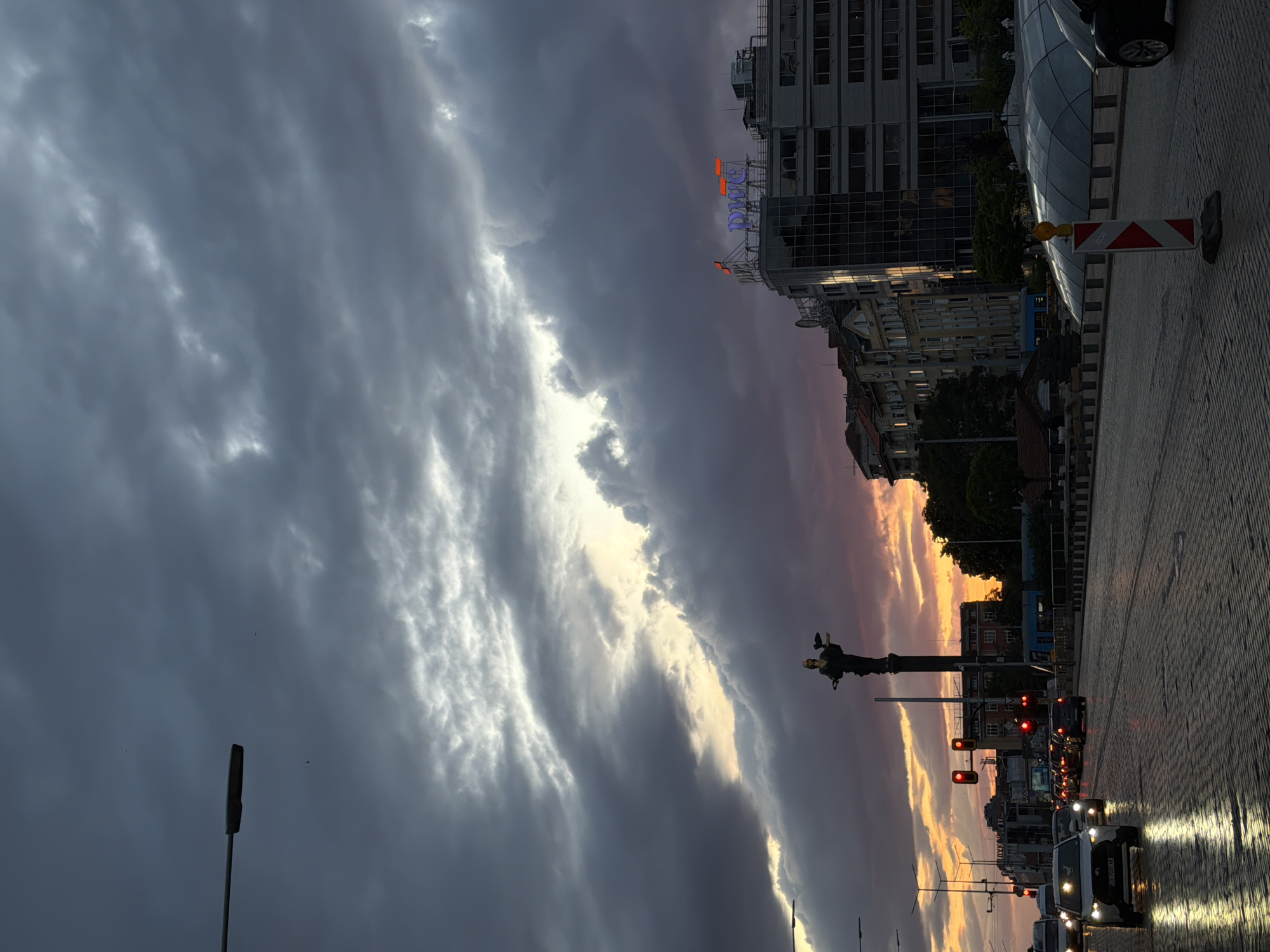 The image depicts a street scene in Sofia, Bulgaria, at dusk, dominated by a dramatic sky. The sky is filled with dark, textured storm clouds on the left, contrasting with a bright, sunlit area near the center, displaying shades of orange and pink towards the horizon. The PwC building is visible on the right side of the image.
A statue atop a tall pillar is centered in the middle ground, facing towards the sunset. Traffic is halted at a red light. Headlights illuminate the cobblestone street in the foreground. Roadside construction equipment is visible on the right side.