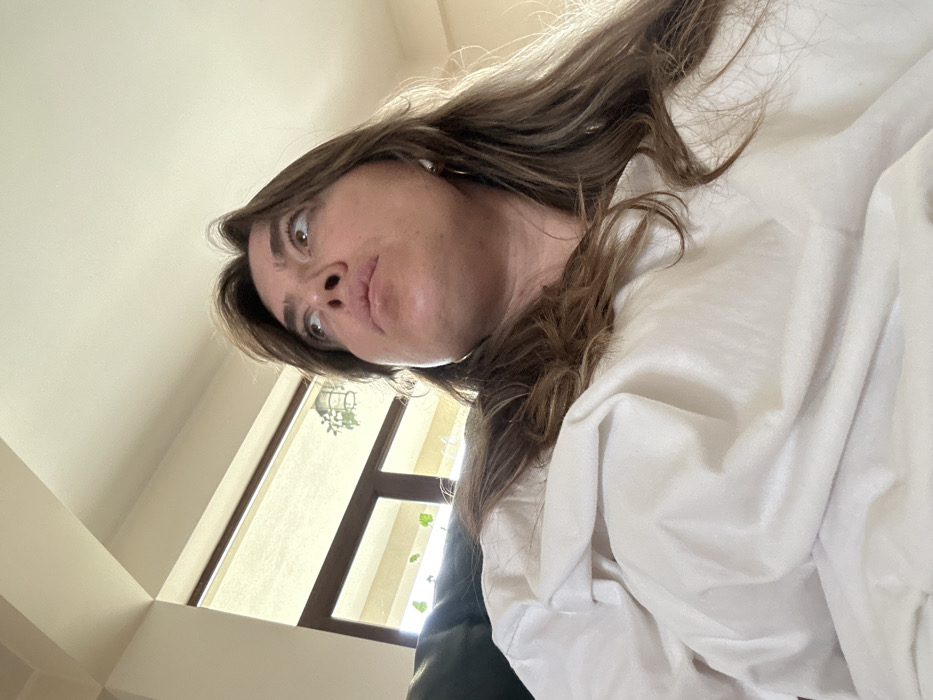 A low-angle, medium close-up shot depicts a female subject positioned indoors in the city of Sighnaghi, Georgia. She possesses shoulder-length, light brown hair with lighter streaks and wears a loose white garment. A white earbud is visible in her right ear. Her head is tilted slightly back, and her gaze is directed upwards and to her right, with a neutral expression. The background features a bright, light-colored ceiling on the upper left. To her left and behind her, a large window or glass door with dark frames allows ambient light into the space. Visible through the window are indistinct green plants, one appearing to be in a hanging basket, and a cream-colored roller blind.