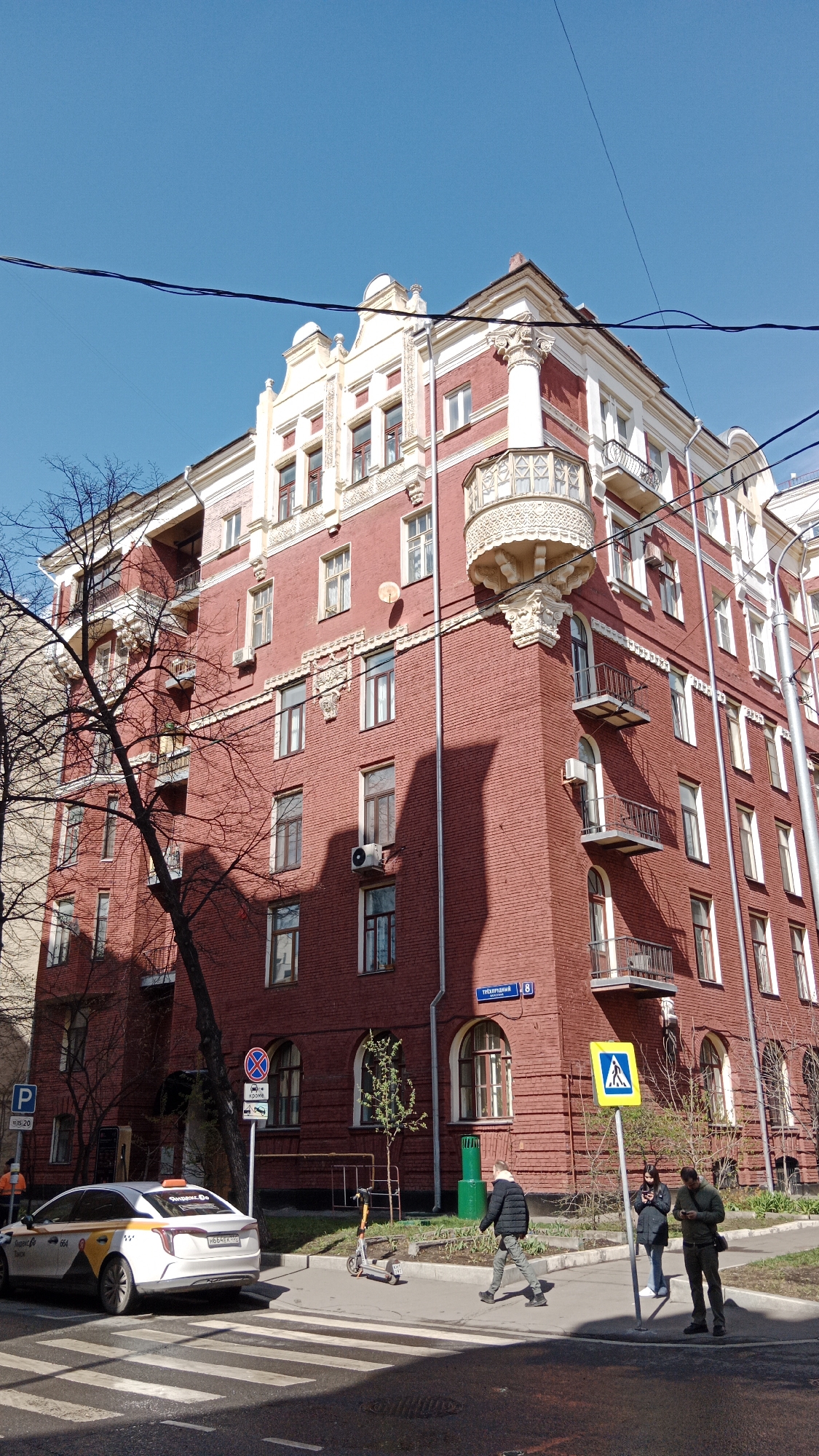A brightly lit street corner in Moscow, Russia, features a distinctive red brick building with elaborate white architectural details under a clear blue sky. The building, likely residential, boasts a prominent rounded bay window with a decorative balustrade on the corner, along with multiple balconies and cornices. White window frames contrast with the red brick facade. Shadows from nearby trees and the building itself fall across its surfaces, indicating a sunny day, likely mid-morning or early afternoon.
On the street level, everyday urban activity unfolds. A white taxi with yellow and grey stripes, bearing the text "Яндекс.Go" (Yandex.Go) on its roof light and license plate H683EE, is parked near a curb. An orange electric scooter stands idle on the sidewalk. Three individuals are visible near a crosswalk: one man walks across it, while two others stand on the sidewalk looking at their mobile phones, positioned next to a yellow pedestrian crossing sign. Another person in an orange vest is partially visible behind the taxi.
Several street signs provide context: a blue "P" parking sign with "5-20" below it, a circular "no stopping" sign (partially obscured), and a blue street sign on the building reading "Трубперский" (Trubpersky) with the number "8", indicating the building address. Overhead power lines crisscross the upper portion of the frame. The trees in the foreground are largely bare, suggesting late winter or early spring.