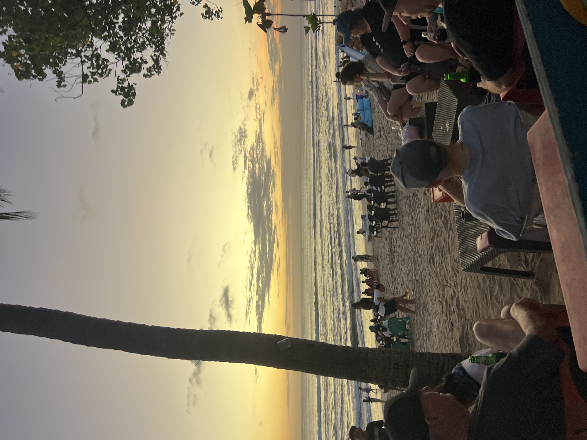 The image depicts a beach scene in Kuta, Indonesia, during sunset. The sky is a gradient of yellow and orange hues, with some clouds scattered across it. In the foreground, several people are seated at tables on the sand, some holding bottles of beer. They appear to be enjoying drinks and conversation. Behind them, the ocean stretches out to the horizon, with gentle waves lapping the shore. Numerous other people are visible on the beach, some walking, others sitting or standing. A tall palm tree stands to the left of the frame, its silhouette dark against the bright sky. In the distance, beach chairs and umbrellas are set up, suggesting a lively atmosphere. The overall impression is one of a relaxed evening on a popular Indonesian beach.