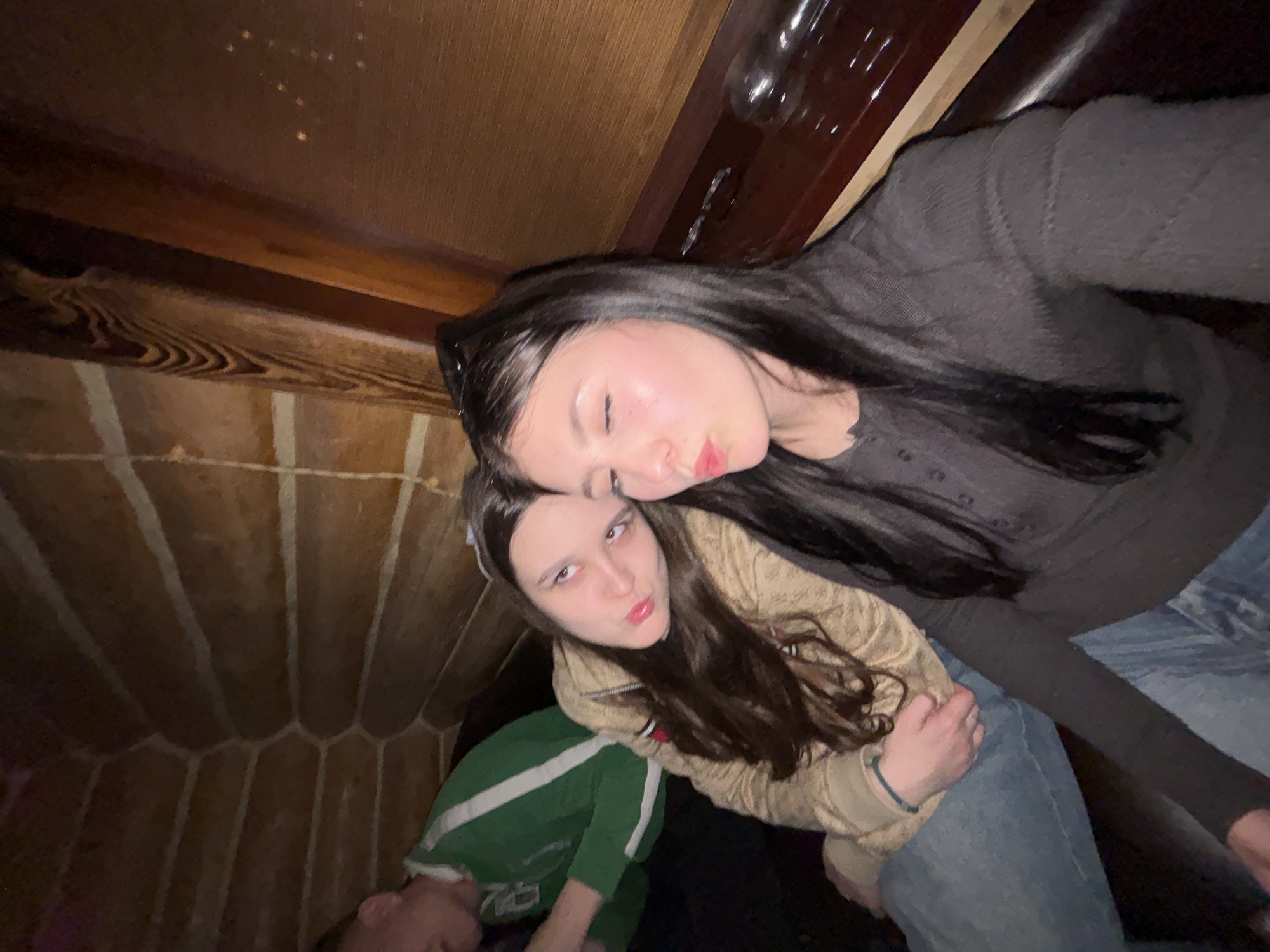 Two young women are posed closely together in a dimly lit interior, likely a room or enclosed space. The woman in the foreground wears a light brown zip-up jacket and green tracksuit jacket, while the other is wearing a dark gray top and jeans. They both have long, dark hair and are making faces towards the camera. The background suggests a rustic or unusual interior, possibly a themed restaurant, with distinctive wood paneling. I cannot determine the location as Chaikovsky, Russia, from the image alone.
