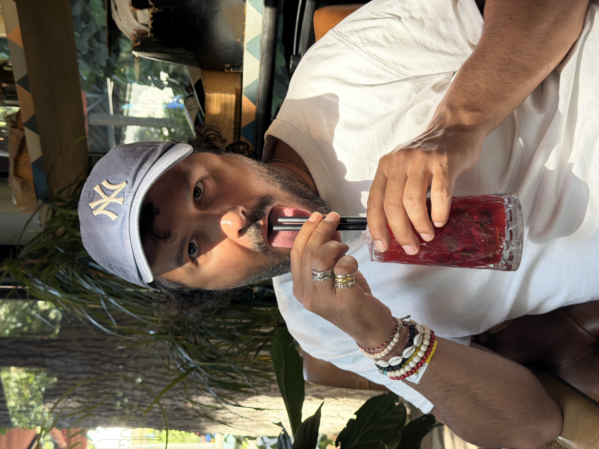 A close-up, slightly rotated, daytime shot shows a man drinking from a glass with two black straws. He is wearing a light-colored New York Yankees baseball cap, a white t-shirt, and several bracelets. He also wears multiple rings on his fingers. The glass contains a red beverage, possibly with fruit or ice. The background includes greenery, suggesting an outdoor or garden-like setting, and hints of furniture and decor. Based on the prompt, it is likely in Tbilisi, Georgia, though the location is not immediately visually identifiable.