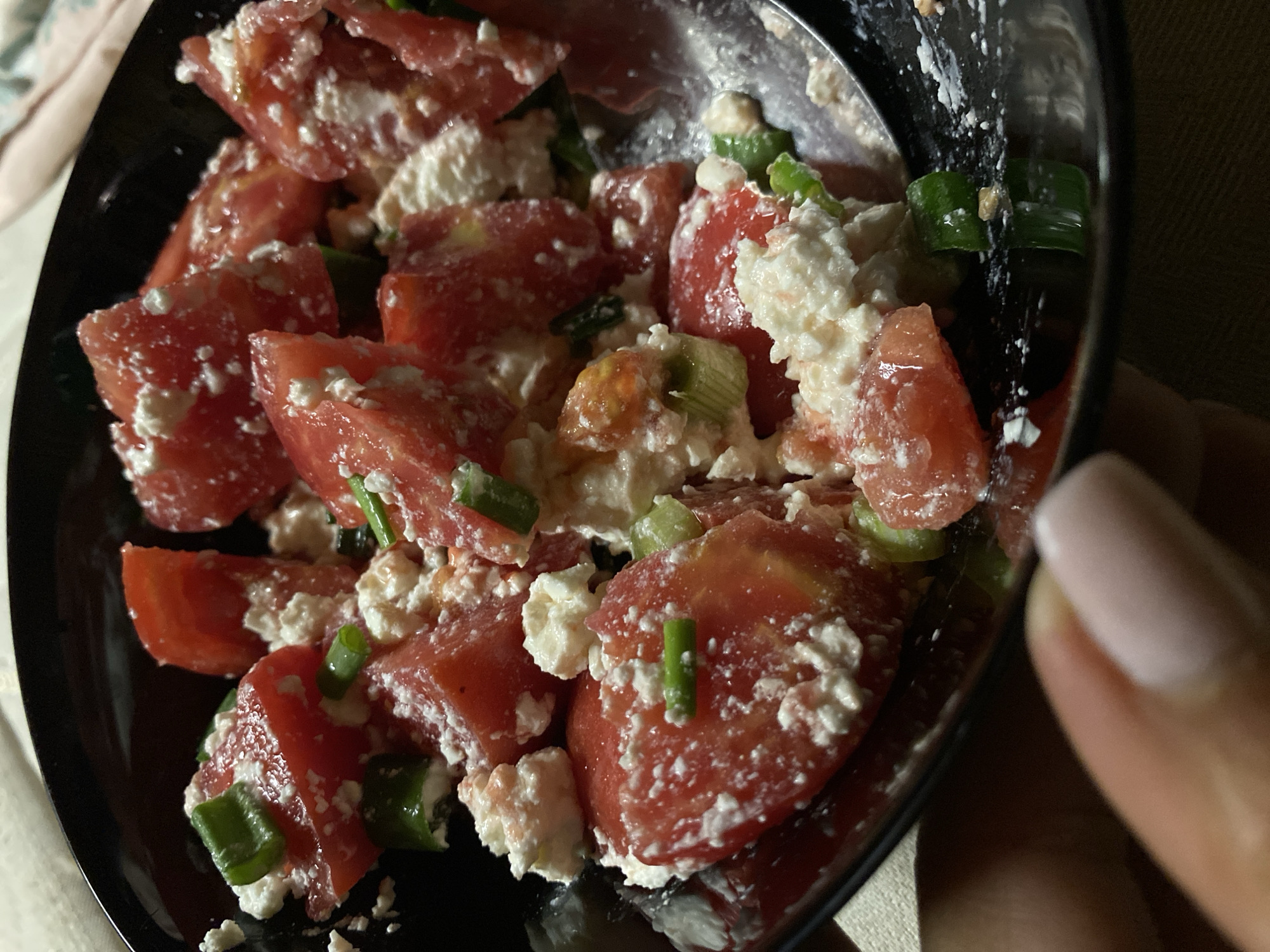 The image shows a close-up of a black bowl filled with a salad. The salad consists of diced tomatoes, crumbled white cheese (possibly feta), and chopped green onions. The bowl is being held by a hand, a fingernail with light pink nail polish is visible. The setting is indoors, possibly a home. The specific location is not discernible from the image.