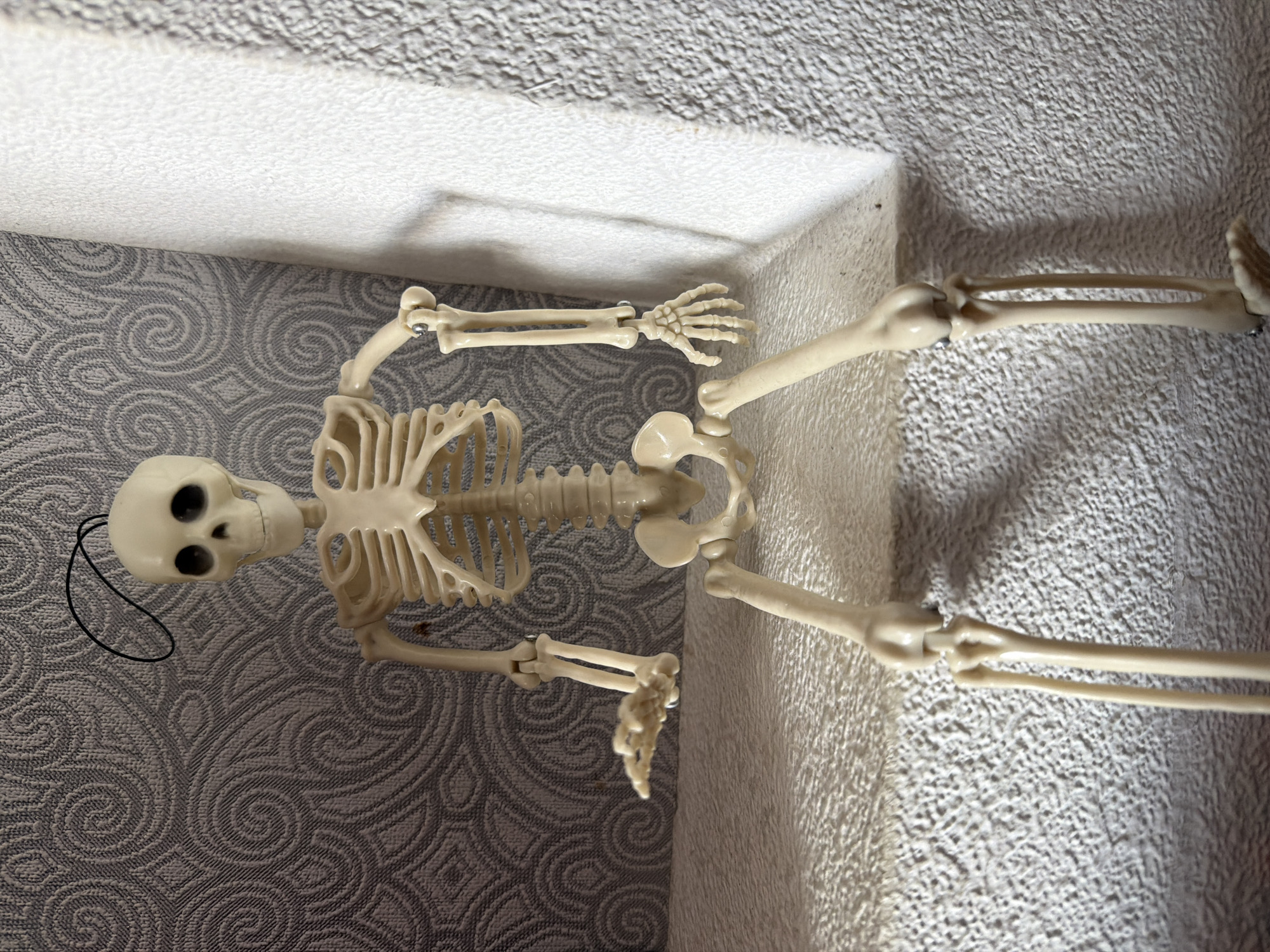 A skeleton figurine is seated on a textured surface, with its legs extended forward. The figurine is positioned in the center of the frame, facing forward. Behind the skeleton, a wall is visible with a grey, swirled pattern. To the right of the skeleton, a white textured surface creates a sharp contrast. The skeleton's left arm is bent at the elbow, with its hand open and palm facing upwards, as if reaching out. Its right arm is also bent, with the hand resting on its leg. The background appears to be an indoor setting, possibly a room or a shelf. No specific location in Baku, Azerbaijan can be identified from the image.