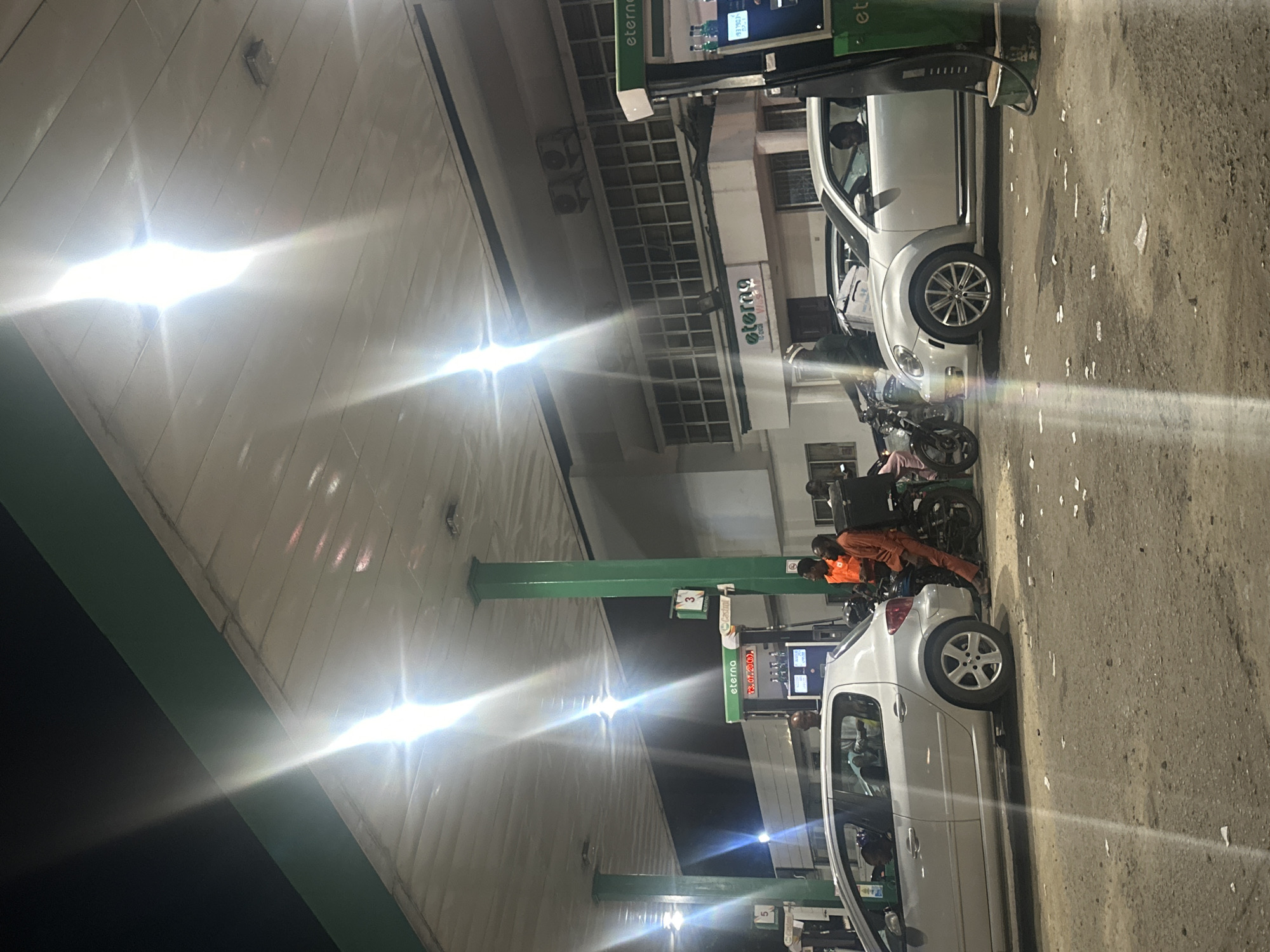 The image shows a nighttime scene at an Eterna filling station in Abuja, Nigeria. Two silver cars are parked at separate fuel pumps. People are gathered around one of the pumps, some wearing orange clothing and sitting on motorcycles. The filling station has a white building with air conditioning units and the Eterna logo. The ground is concrete. Bright overhead lights illuminate the scene.