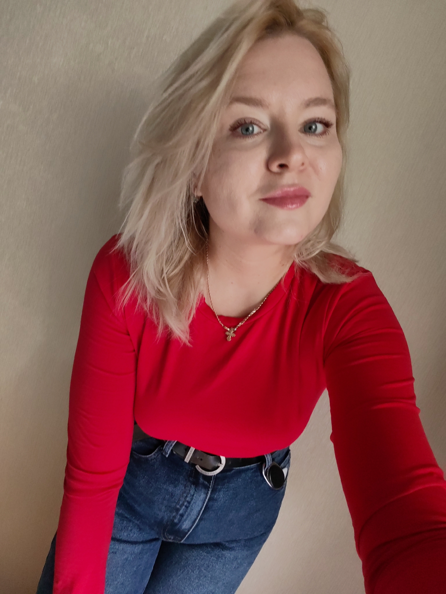 The image shows a medium-shot selfie of a blonde woman with blue eyes, wearing a red long-sleeved shirt and blue jeans. She is leaning forward, with her head slightly tilted. She has a light-colored necklace with a small pendant. Her hair is light blonde and appears somewhat messy. The background is a plain, textured light-colored wall. The image does not provide any information or visual cues to determine the specific location within the city of Izhevsk, Russia, or any other city.