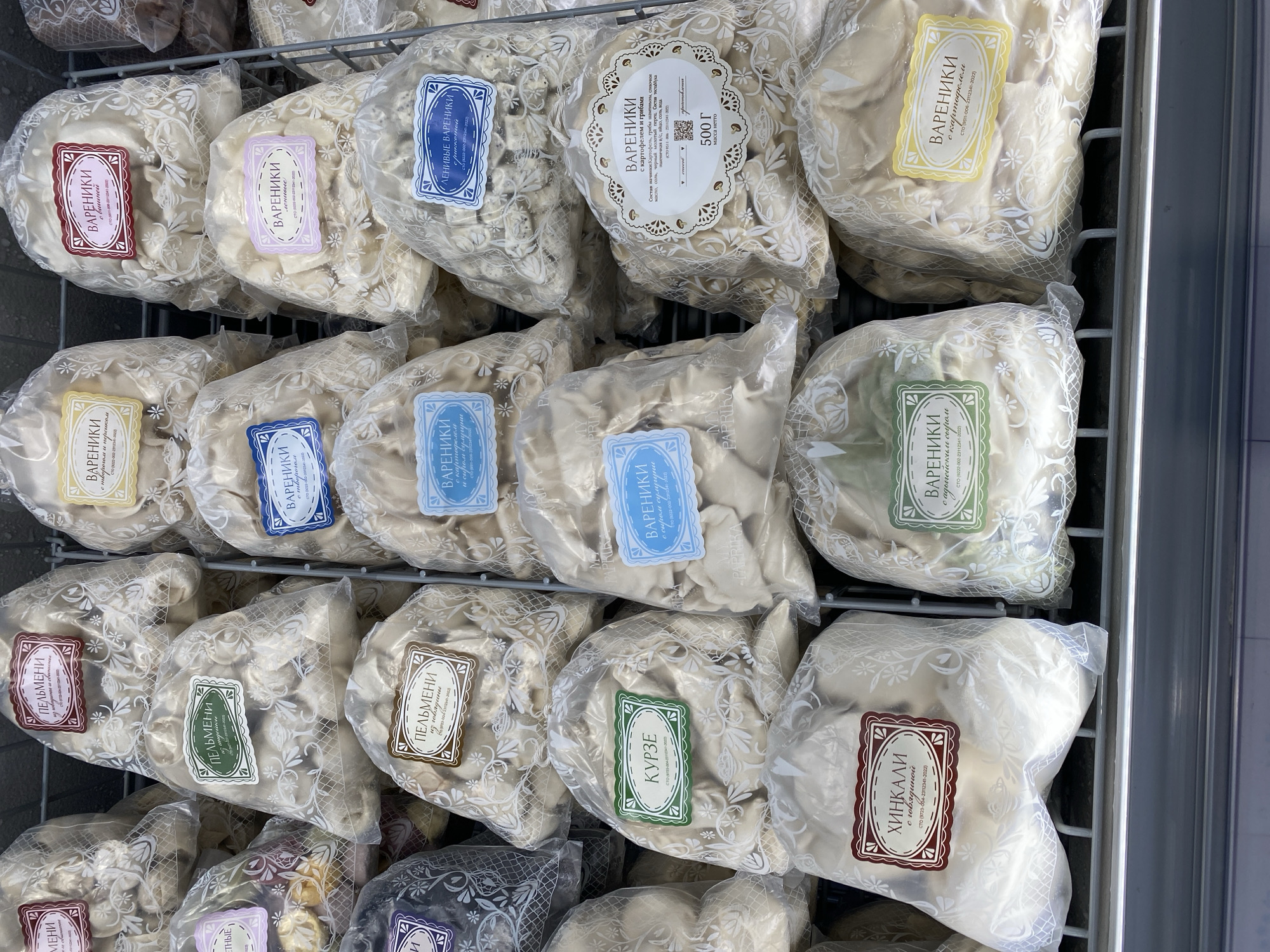 The image displays a refrigerated display case filled with various frozen dumplings and pelmeni, packaged in transparent plastic bags. The bags are adorned with colorful labels indicating the product type, such as "Пельмени" (Pelmeni), "Вареники" (Vareniki), and "Хинкали" (Khinkali), along with specific fillings and ingredients. The packaging features floral patterns and decorative borders. The products are arranged in multiple rows, filling the display case. The background is a metal shelving unit. The scene is likely in a retail setting, possibly a grocery store or supermarket in Novaya Usman', Russia, given the context. There are no people or animals visible in the image.