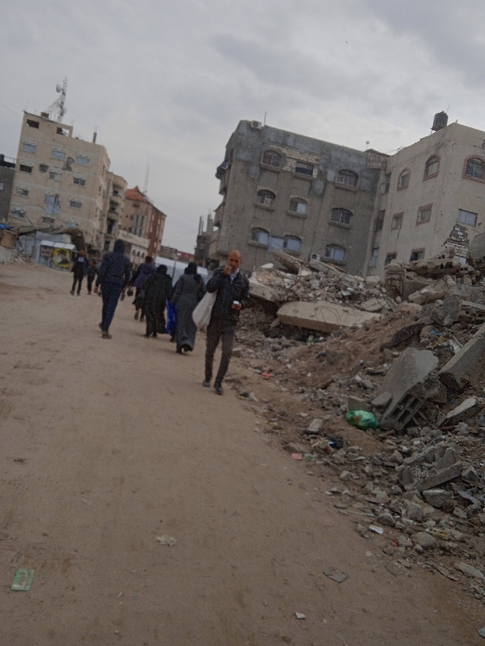 The image depicts an urban street scene in Khan Yunis, Palestinian Territory, under an overcast sky. An unpaved dirt road stretches across the foreground and midground. In the foreground, slightly right of center, an adult male with a bald head or close-shaven hair and a short beard walks towards the viewer. He wears a dark jacket, dark trousers, and light-colored shoes, holding a dark object in his right hand and with his left hand near his mouth. A white bag is draped over his left shoulder. Behind him, on the left side of the road, a group of approximately six to seven individuals, mostly dressed in dark clothing, walks away from the viewer; one person in this group carries a blue bag. To the right of the road, a large accumulation of building rubble, consisting of concrete slabs, broken bricks, and exposed rebar, forms a mound, with a green plastic bag visible on its surface. In the background, several multi-story concrete buildings are visible; the building on the far left features a communication tower on its roof, while the structure in the center-right background displays significant damage with exposed rebar and structural elements.