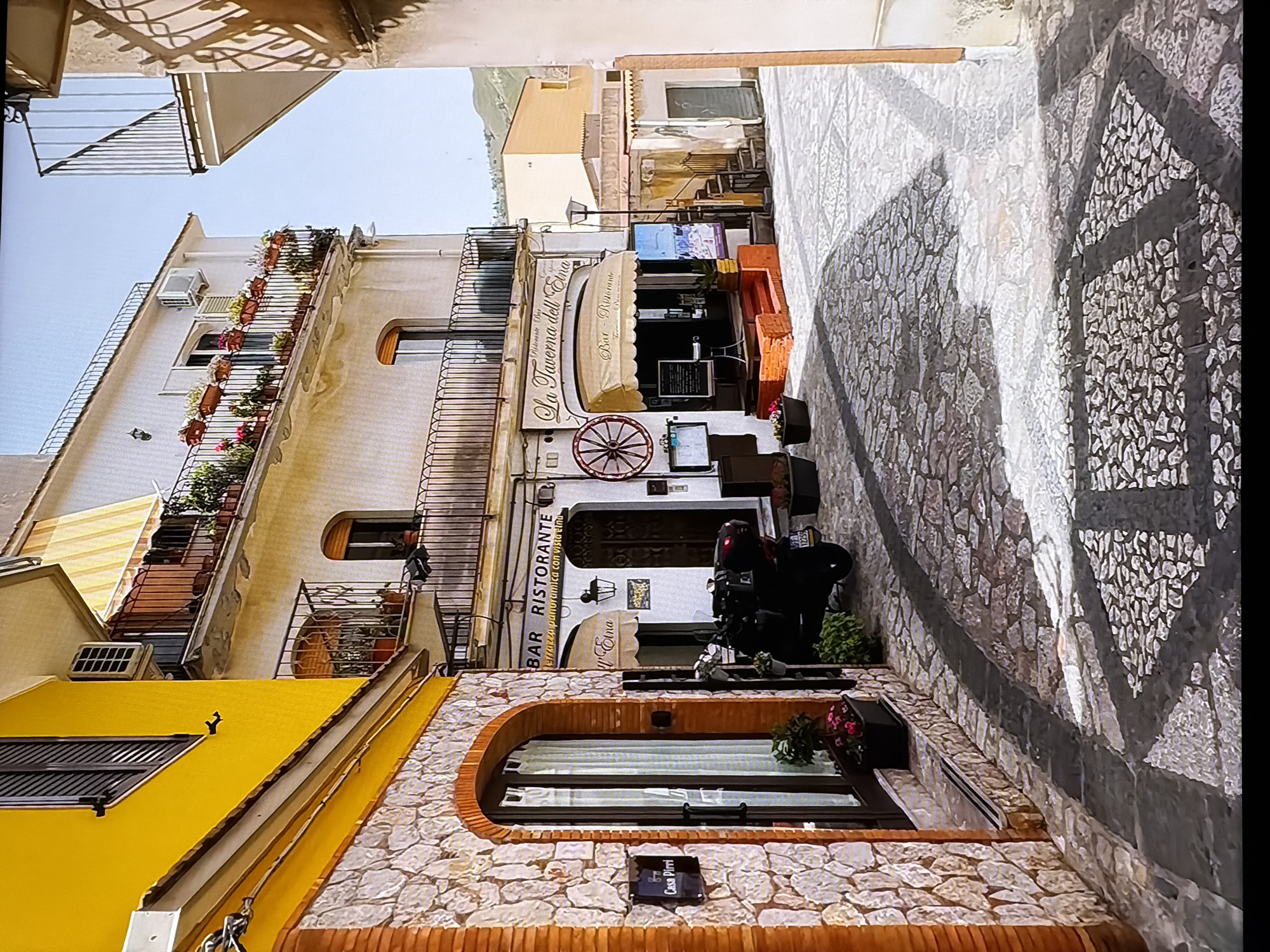 The image depicts a narrow, cobblestone street lined with buildings. On the left, a building with a bright yellow facade features a stone-clad lower level with an arched entryway. Above the entryway, a sign reads "Casa Pirri". Adjacent to this building is a white building with balconies adorned with potted plants. On the right side of the street, a restaurant named "La Taverna dell'Etna" is visible, with an awning, tables, and menus. A dark-colored scooter is parked in front of the restaurant. Shadows from the buildings and a street lamp are cast across the cobblestone pavement, indicating it is daytime. The architectural style suggests a Mediterranean setting, likely in Italy. There is no visible indication of the location being Chirchik, Uzbekistan.