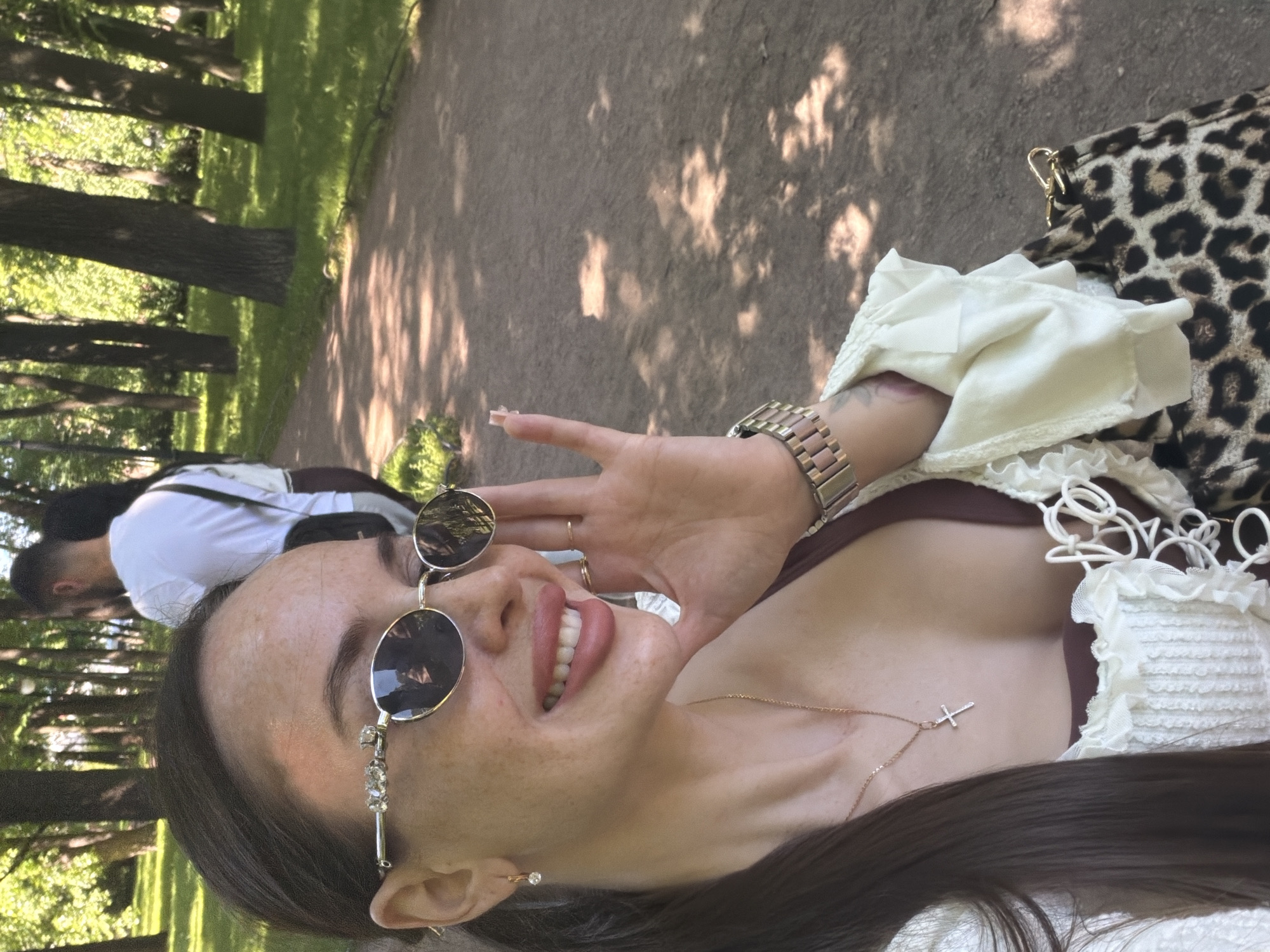 A woman is depicted in a close-up outdoor shot, smiling and looking to her right, in a park located in Saint Petersburg, Russia. She has long, dark brown hair pulled back, a fair complexion with numerous freckles visible on her forehead, nose, and cheeks. She wears round, dark-lensed sunglasses with decorative clear stones on the left temple. Her lips are full and colored with a pinkish-mauve lipstick, revealing white teeth in a broad smile. She wears a small gold hoop earring with a clear stone in her visible left ear, a thin gold chain necklace with a small, stone-embellished cross pendant, and a gold-toned metal link watch on her left wrist. Her attire includes an off-white or cream-colored top with ruffled trim and front lacing or decorative ties, layered over a darker, possibly brown, garment. Her right hand is raised, with fingers lightly touching her sunglasses and cheek. A brown and black leopard-print handbag with gold hardware is visible on her lower right side. The background features a wide, unpaved path winding through a park with lush green grass and numerous mature trees with dark trunks and green foliage. Dappled sunlight creates varied shadow patterns on the path. Two individuals are walking away from the camera on the path in the middle ground, out of focus; the closer person is a bearded man wearing a white t-shirt and a dark cross-body bag.